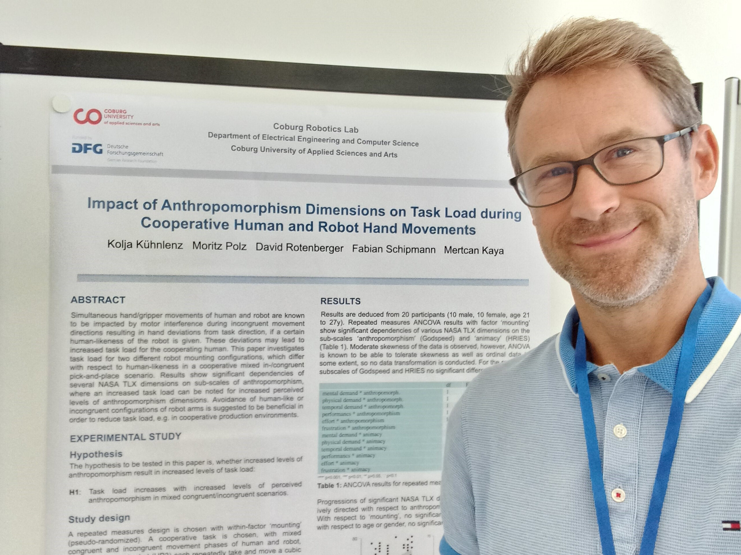 Eine Person mit Brille steht neben einem wissenschaftlichen Poster mit dem Titel „Einfluss anthropomorphistischer Dimensionen auf die Aufgabenbelastung bei kooperativen menschlichen und Roboterhandbewegungen“ in einem Robotiklabor der Hochschule Coburg. Das Poster enthält Abschnitte zu Zusammenfassung, Studie und Ergebnissen.