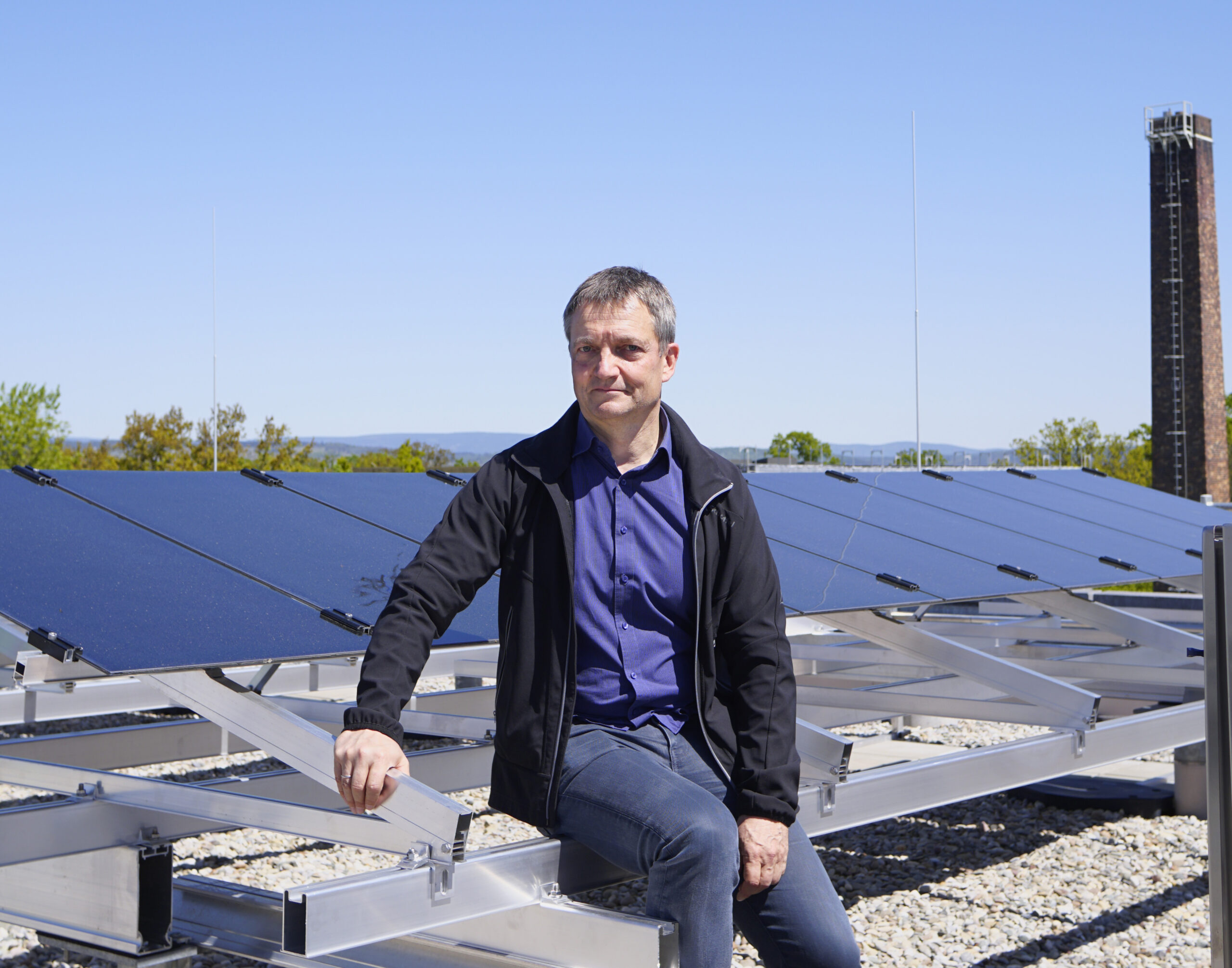 Eine Person sitzt auf einer Metallkonstruktion neben Solarmodulen unter einem klaren blauen Himmel, vermutlich an der Hochschule Coburg. Sie trägt eine schwarze Jacke und ein blaues Hemd. Im Hintergrund sind Bäume und ein Schornstein zu sehen.