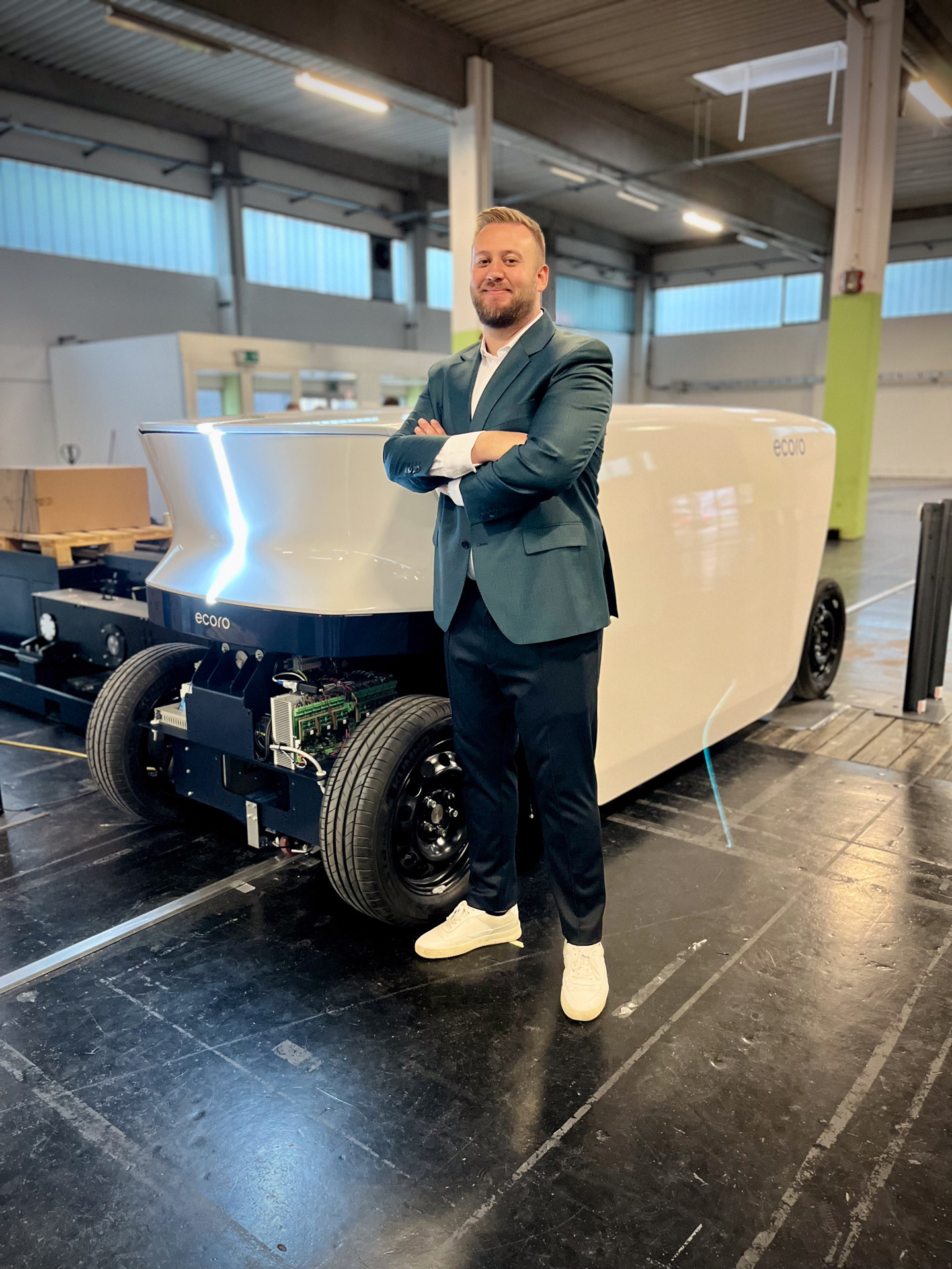 A person stands confidently with crossed arms next to a sleek, futuristic white vehicle prototype labeled "ecoro" in a spacious industrial setting at Hochschule Coburg. The vehicle boasts a minimalistic design, while the person is stylishly dressed in a suit and sneakers.