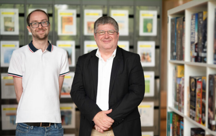 Two men stand smiling in a library-like setting at Hochschule Coburg. The man on the left wears a white polo shirt and glasses, while the man on the right sports a black blazer over a white shirt. Shelves filled with books and magazines form the perfect backdrop to this intellectual scene.