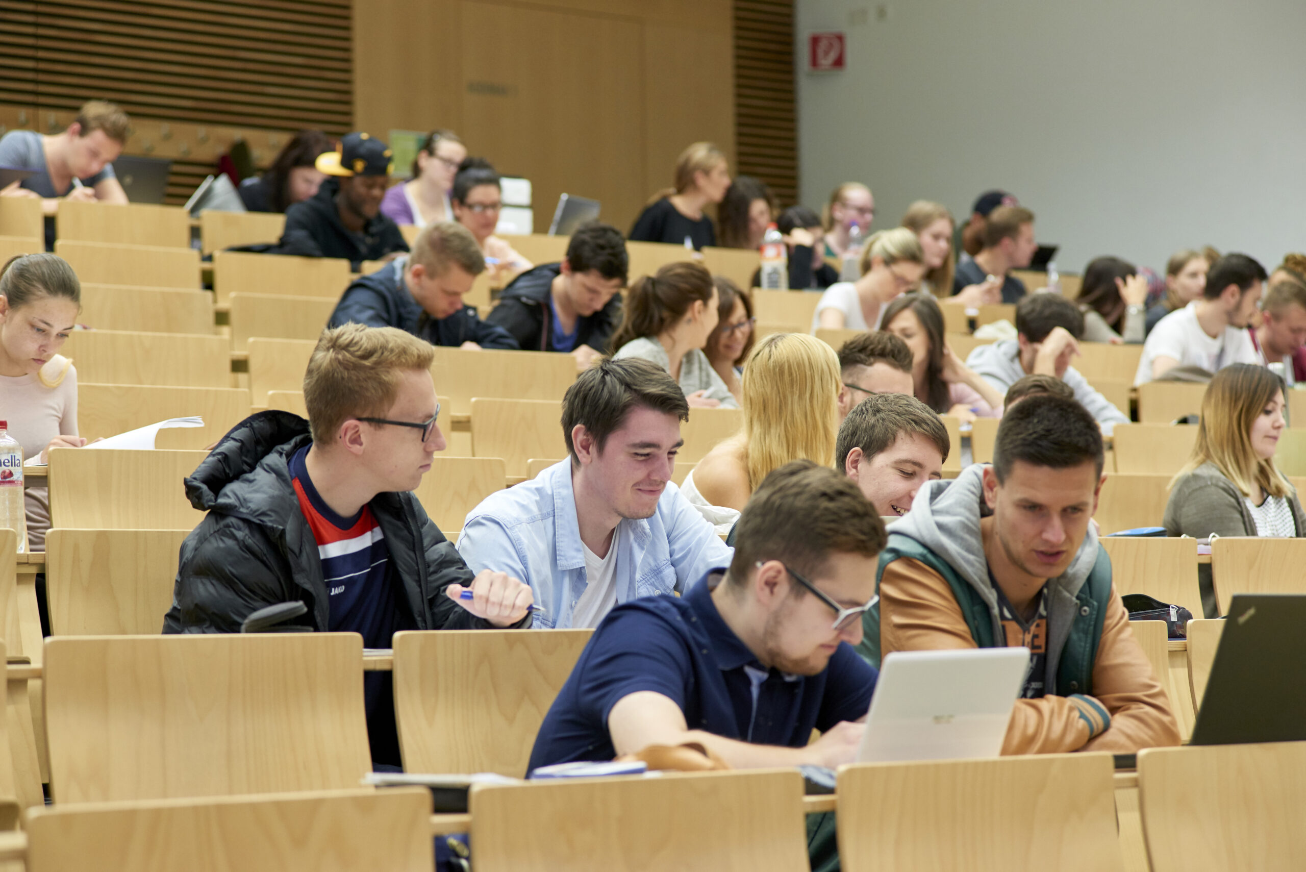 Im modernen Hörsaal der Hochschule Coburg sitzt eine große Gruppe von Studierenden aufmerksam. Viele machen sich Notizen oder benutzen Laptops und sind voll und ganz mit dem präsentierten Stoff beschäftigt. Der Raum kombiniert elegante Holzsitze mit modernem Design und fördert so eine Atmosphäre der Konzentration und des Lernens.