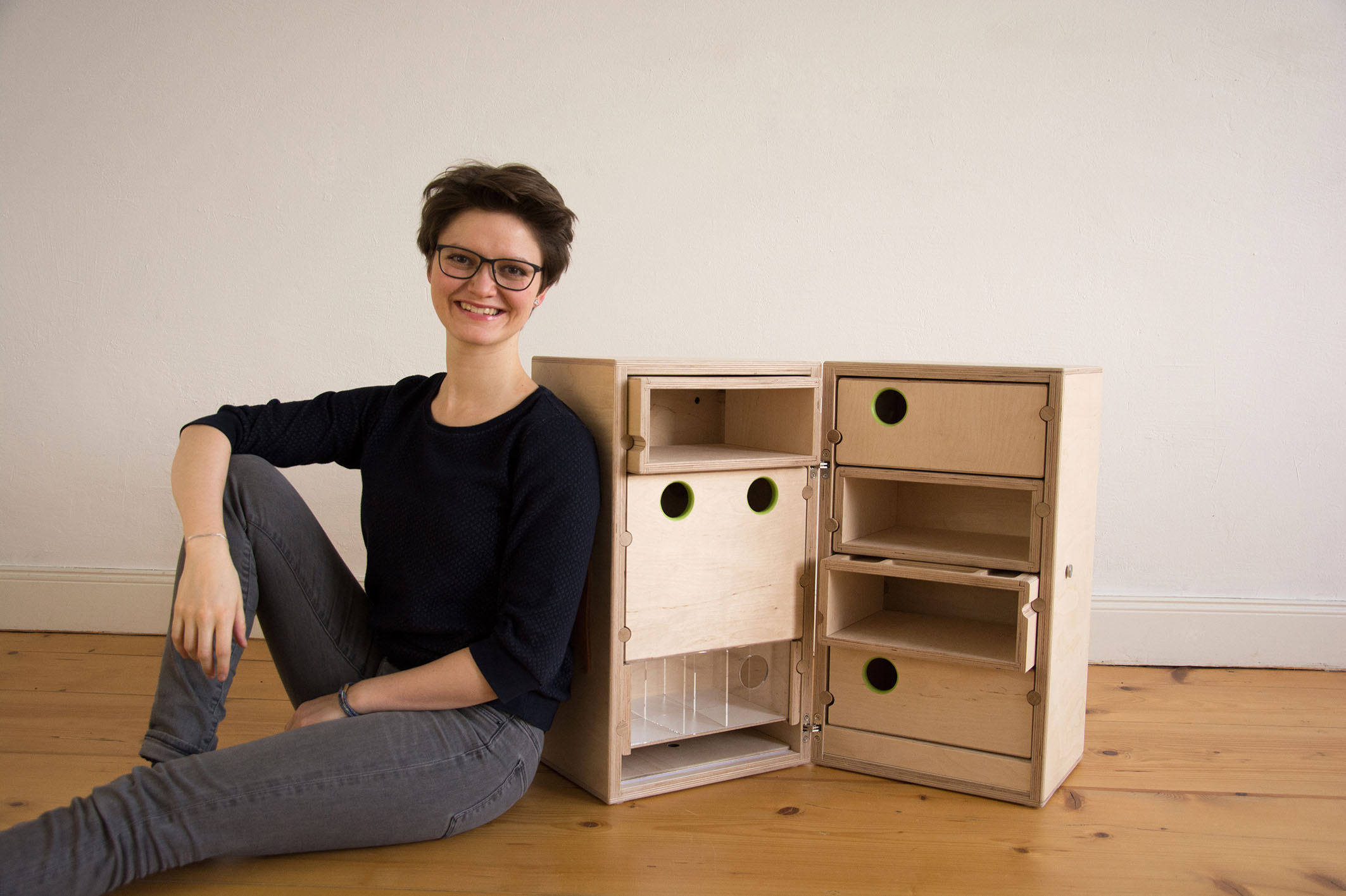 Eine Person mit kurzen Haaren und Brille sitzt auf einem Holzboden neben einem einzigartigen Schrank, der mit kreisrunden grünen Löchern verziert ist. Die offenen Fächer des Schranks offenbaren sein faszinierendes Design, das an den kreativen Geist der Hochschule Coburg erinnert. Eine schlichte, helle Wand vervollständigt die ruhige Szene.