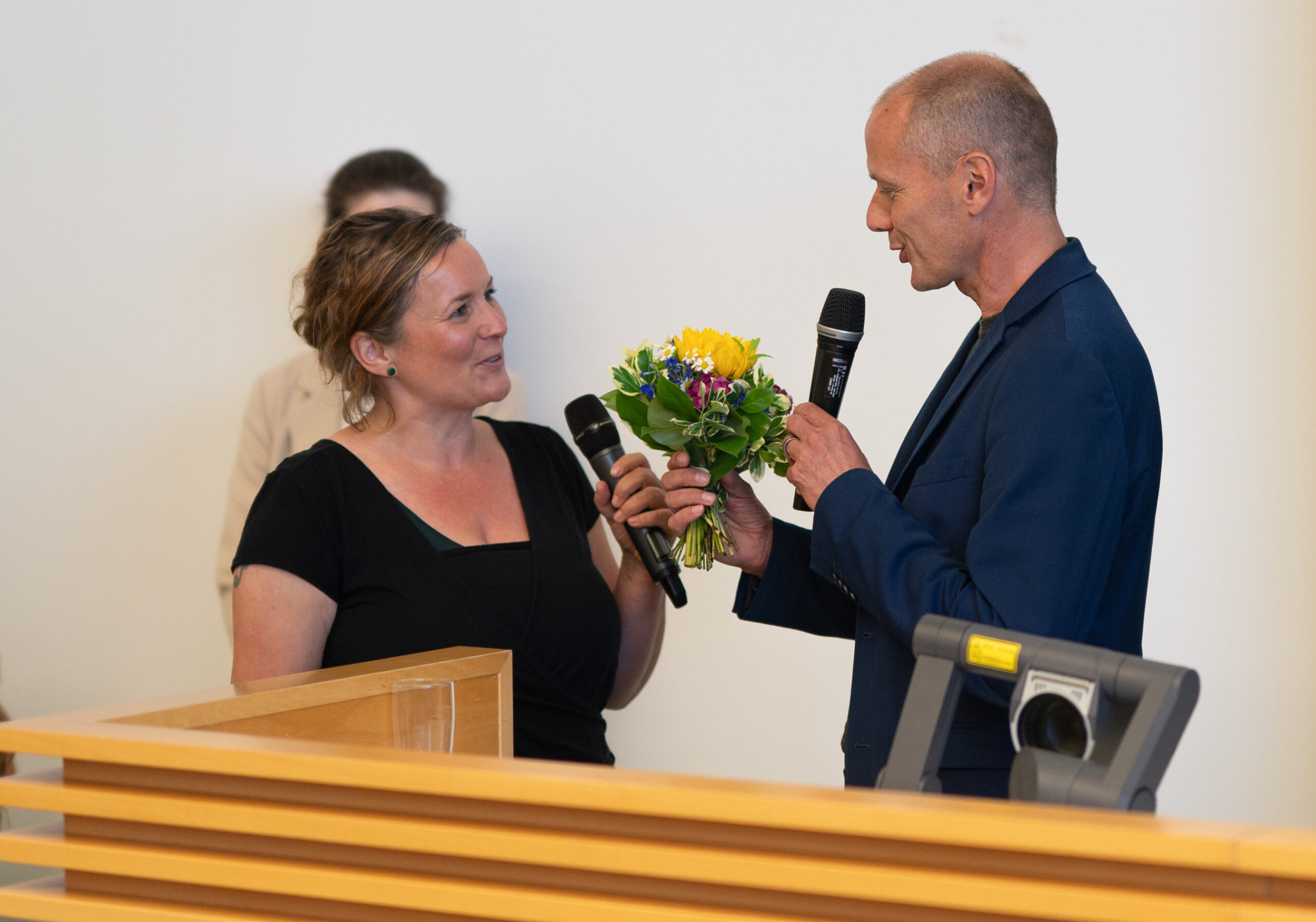 A man and a woman stand together, each holding a microphone, with the woman also cradling a small bouquet of flowers. They seem to be speaking at an event for Hochschule Coburg, with a wooden lectern and a projector in the foreground.