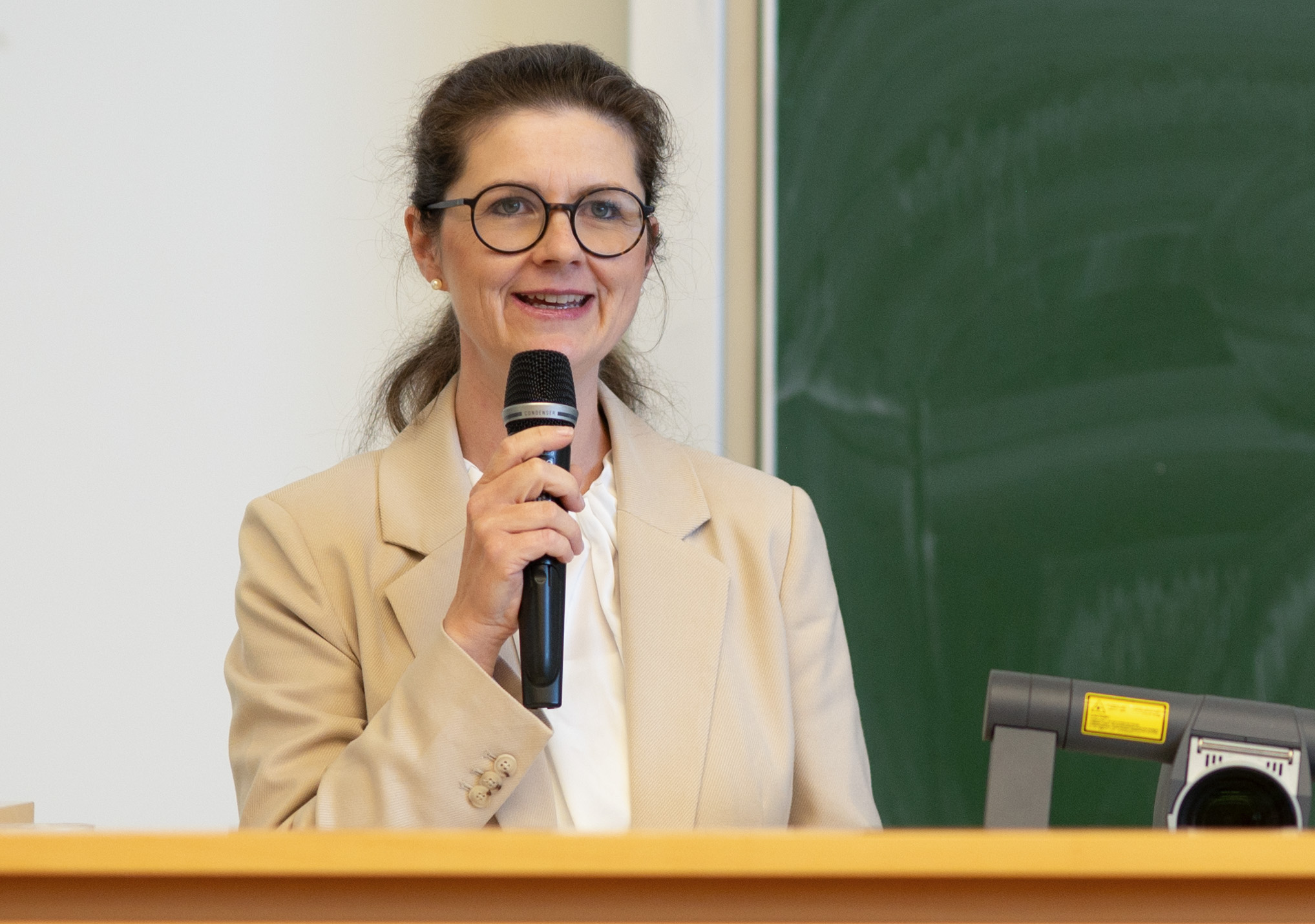 A woman with glasses and a beige blazer is speaking into a microphone while standing at a podium. She is in front of a green chalkboard, addressing students in what appears to be a Hochschule Coburg classroom or lecture hall.