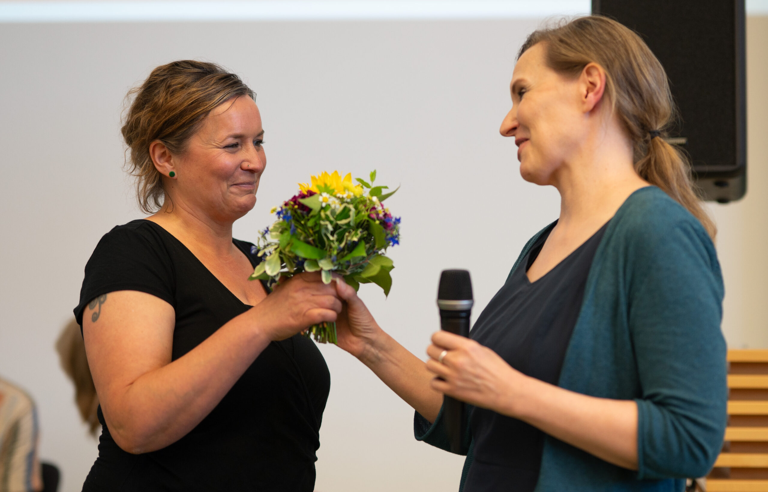 Two women are standing indoors at Hochschule Coburg. One woman hands a colorful bouquet of flowers to the other, who is holding a microphone. Both are smiling warmly, and the atmosphere appears celebratory.