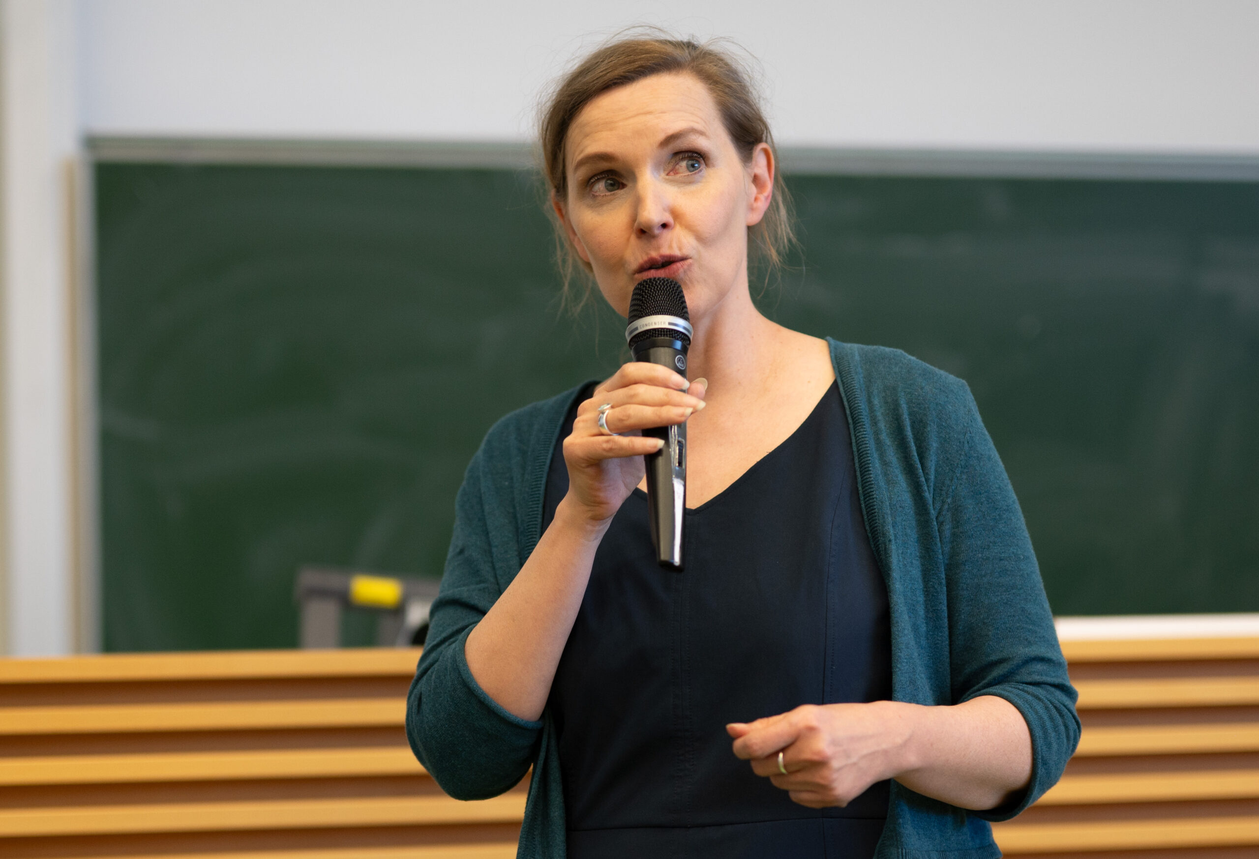 A woman is speaking into a microphone at Hochschule Coburg, standing in front of a chalkboard. She is wearing a dark blue dress and a green cardigan, with her hair tied back.