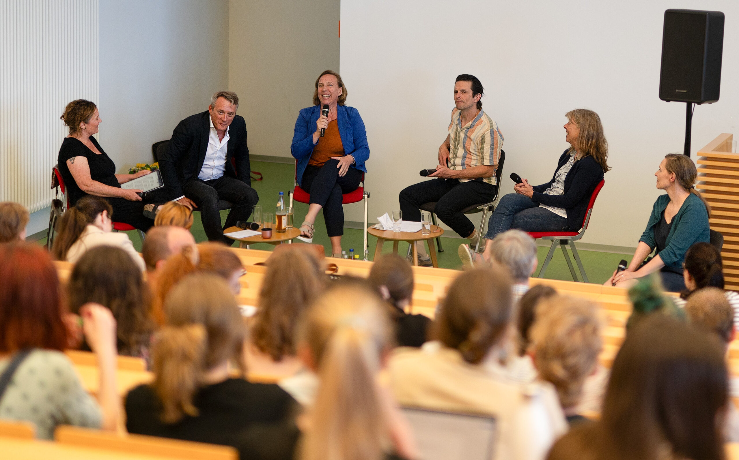 A group of six people sits in chairs on a stage at Hochschule Coburg, engaging in a panel discussion. They are surrounded by an audience in the lecture hall. Some participants hold microphones, and there are drinks on the small tables in front of them.