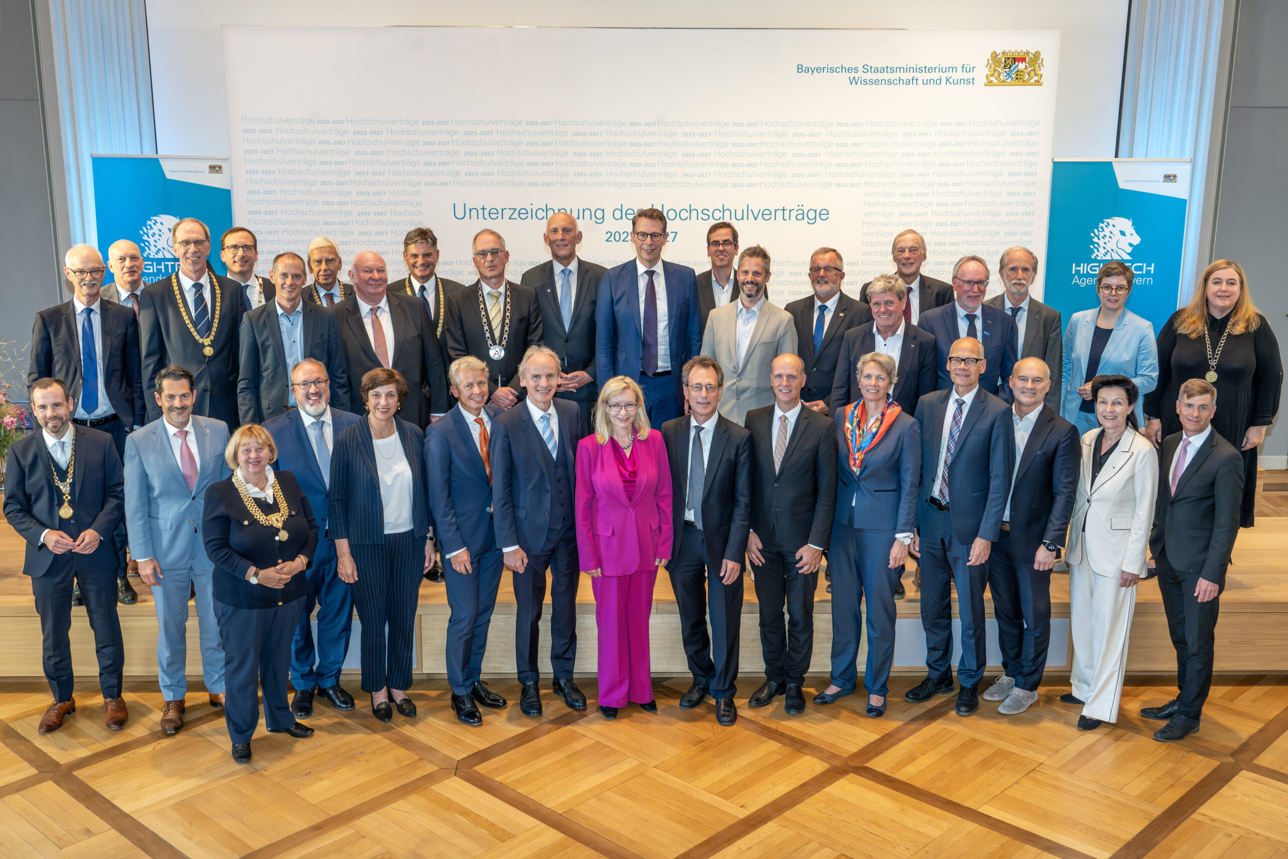 Eine große Gruppe professionell gekleideter Männer und Frauen der Hochschule Coburg steht auf einer abgestuften Plattform und posiert für ein Foto in einem Konferenzrahmen mit der Aufschrift „Unterzeichnung der Hochschulverträge“. Sie sind von blauen Bannern mit Text umgeben.