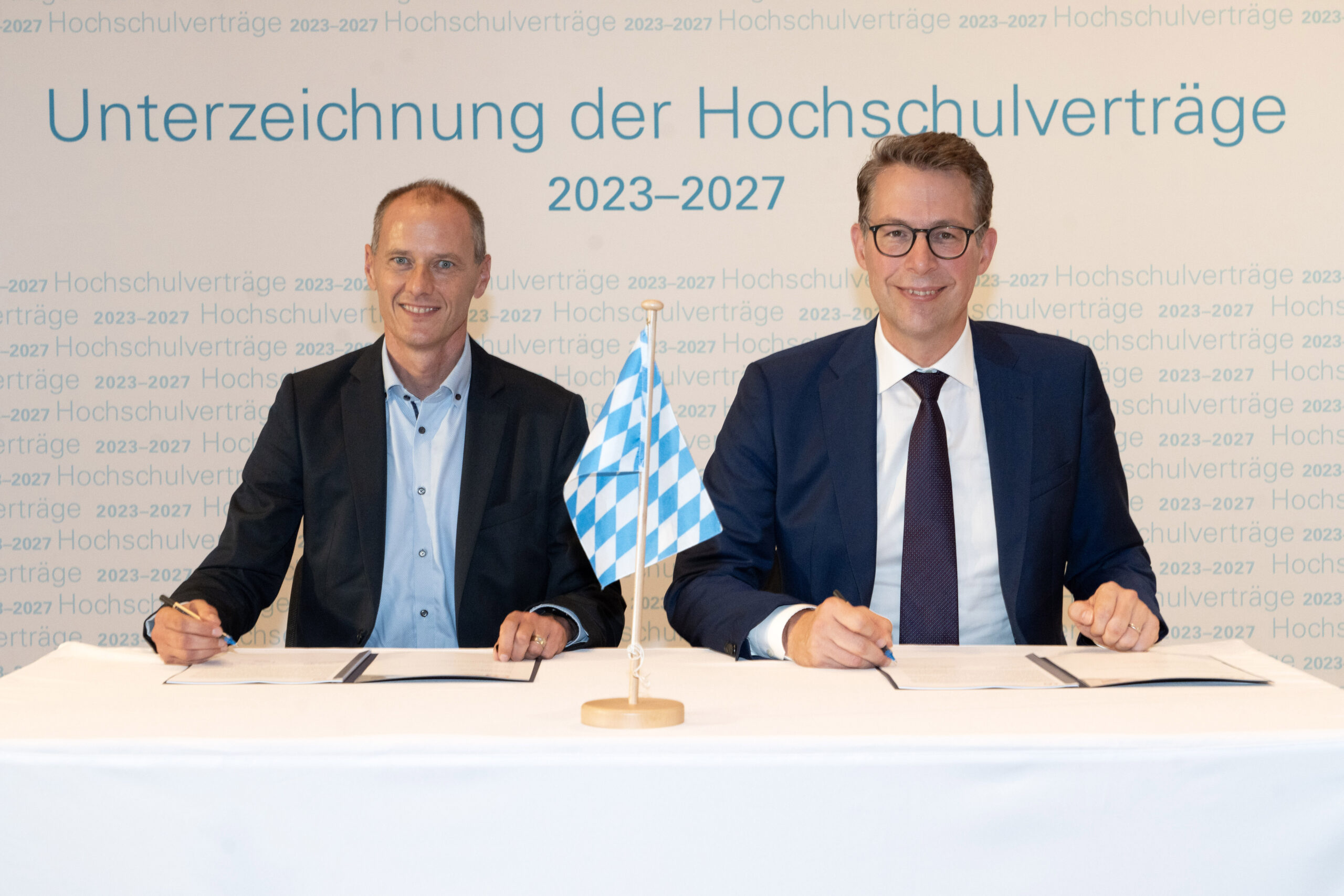 Zwei Männer sitzen lächelnd an einem Tisch mit Dokumenten und unterschreiben Verträge unter dem Banner „Unterzeichnung der Hochschulverträge 2023–2027“. Zwischen ihnen hängt eine bayerische Fahne – ein Bekenntnis zur Hochschule Coburg.