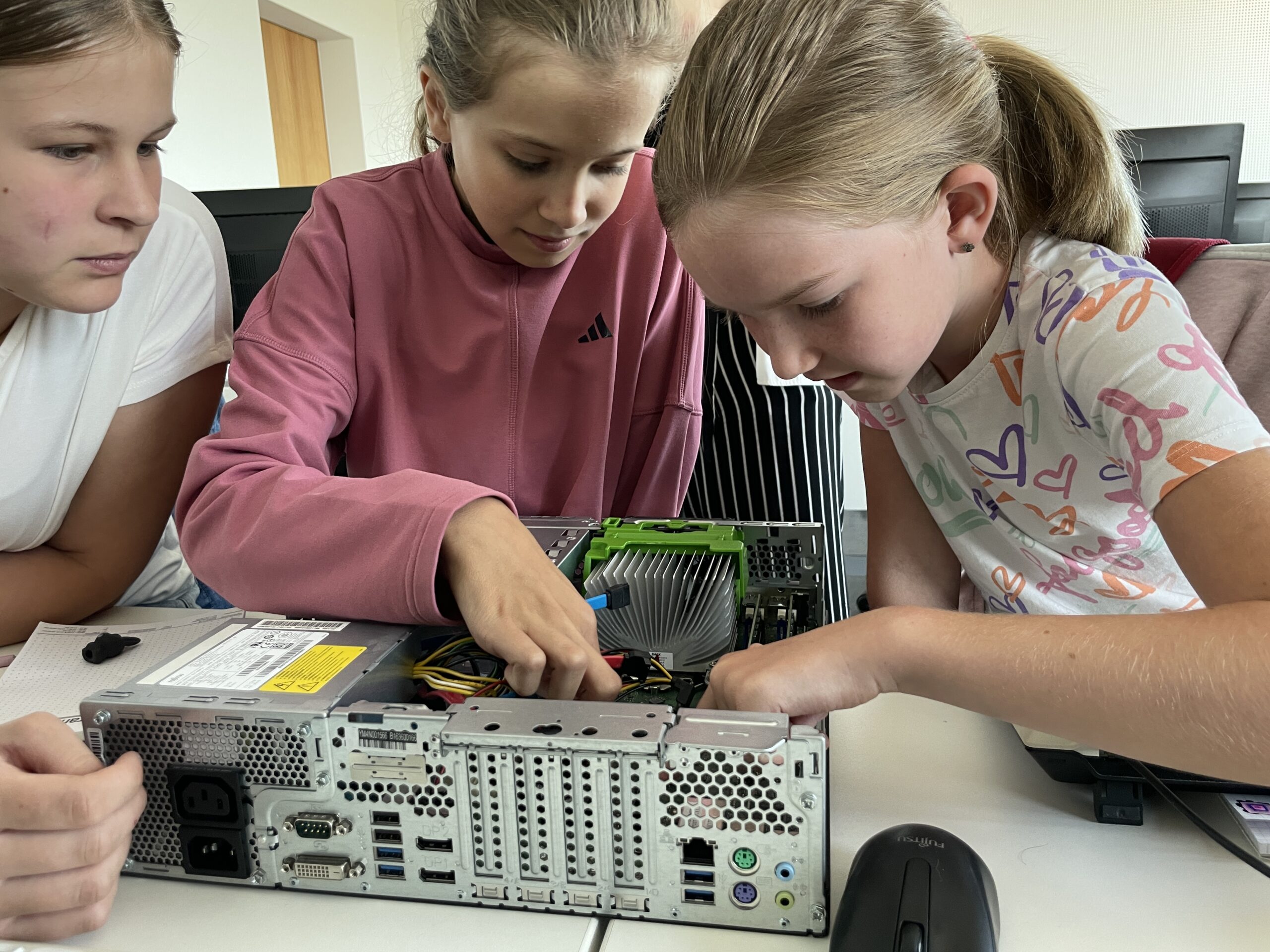 Three girls from Hochschule Coburg are working together on a computer assembly. They're focused on connecting components inside an open computer case on a table. One points inside, guiding the others, while a mouse and papers lie nearby.