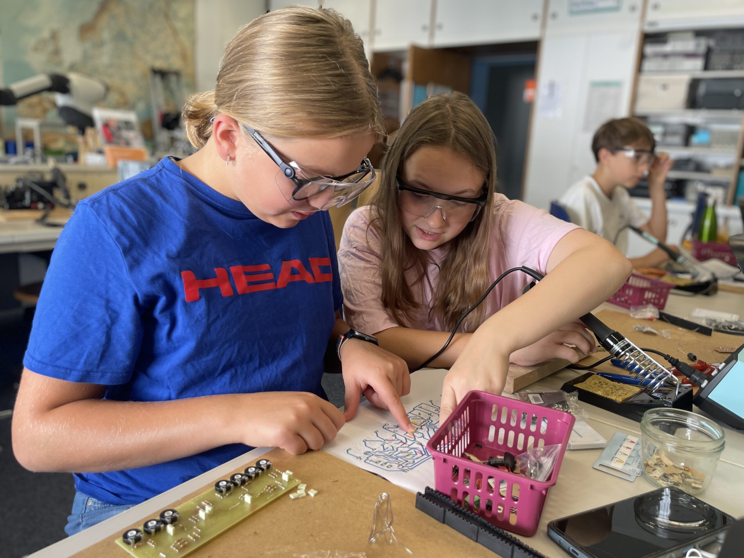 In a classroom at Hochschule Coburg, two girls with safety glasses focus intently on an electronics project, analyzing a circuit board diagram. Another student is in the background, similarly engrossed in their work. Tables are scattered with various electronic components.