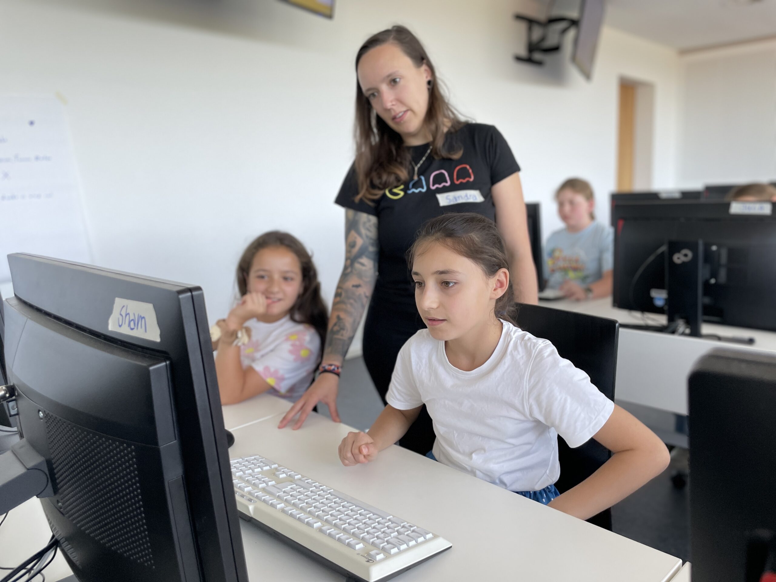 In a classroom setting at Hochschule Coburg, a woman assists two young girls who are seated and working on a computer. The girls are focused on the screen as she observes, providing guidance. Other students and computers fill the background, creating an atmosphere of collaborative learning.