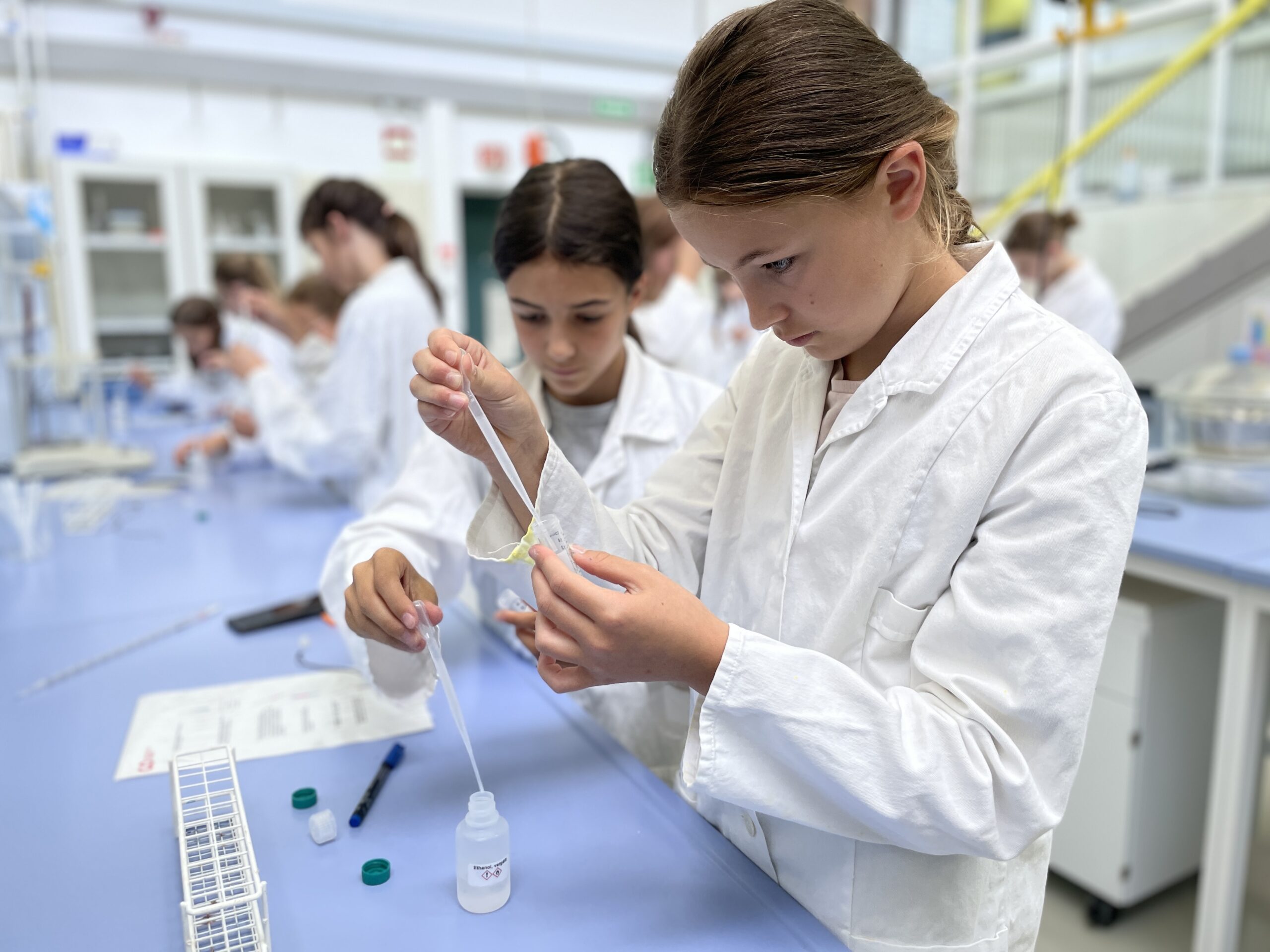 Students in white lab coats from Hochschule Coburg conduct an experiment in the science lab. They use droppers and focus intently on their tasks. The room is equipped with blue tables, lab equipment, and visible safety protocols.