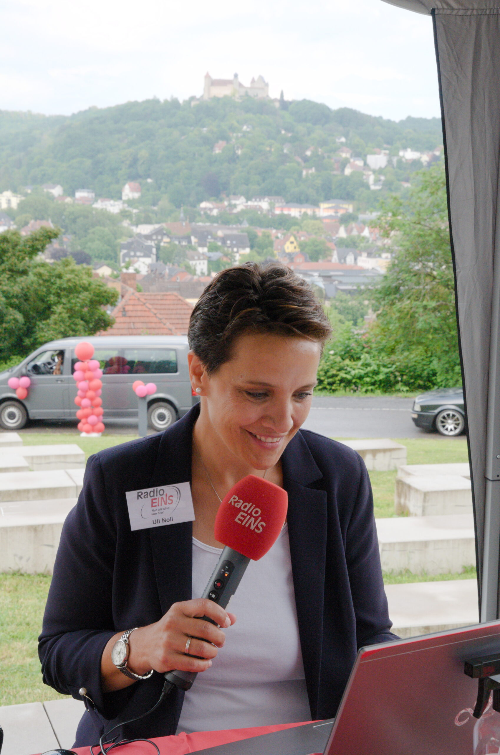 Eine Person mit kurzen Haaren hält ein rotes „RadioEINS“-Mikrofon und lächelt in einen Laptop. Sie sitzt unter einem Vordach vor einer Burg auf einem Hügel, neben einem grauen Lieferwagen, der mit rosa Luftballons geschmückt ist. Auf ihrer Jacke prangt dezent das Emblem der Hochschule Coburg.