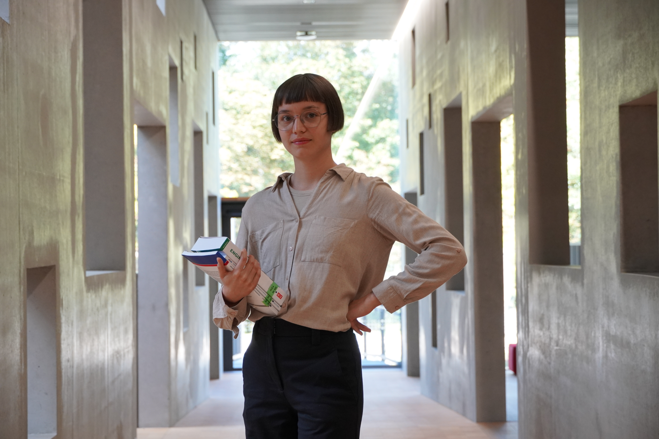 Eine Person mit kurzen Haaren und Brille steht in einem Flur der Hochschule Coburg und hält mehrere Bücher und Papiere in der Hand. Der Flur ist modern gestaltet und hat auf beiden Seiten symmetrische Öffnungen, während am Ende Sonnenlicht durch das Glas fällt.