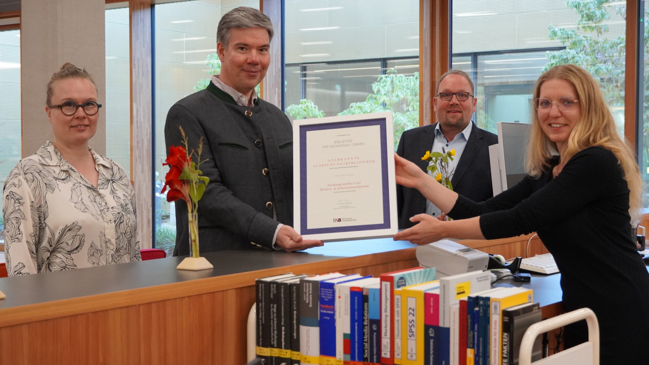 Vier Personen stehen in einem Büro der Hochschule Coburg. Eine Frau überreicht einem Mann ein gerahmtes Zertifikat, während zwei andere lächelnd daneben stehen. Im Vordergrund ist eine Büchersammlung zu sehen.