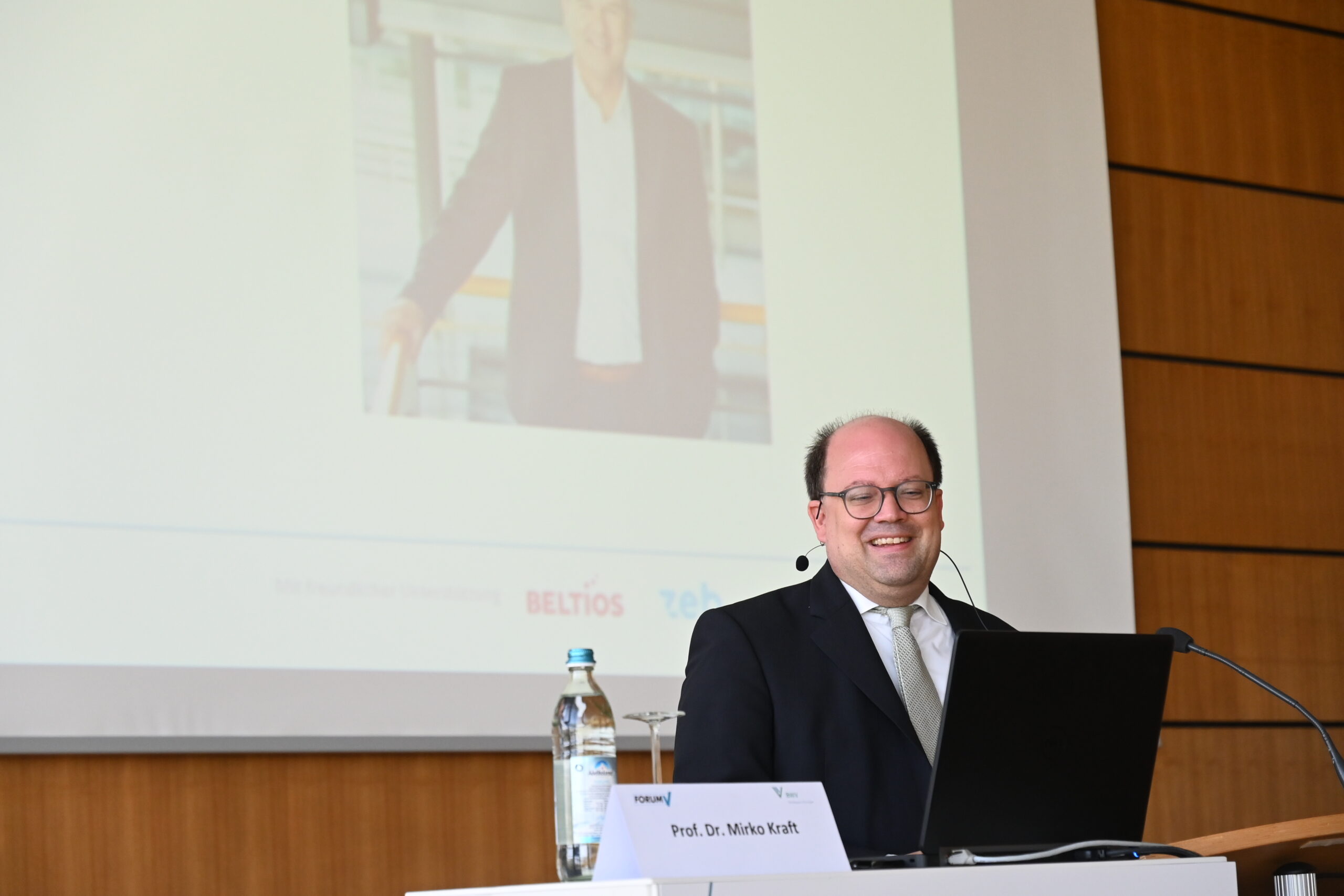 Eine Person im Anzug steht mit einem Laptop an einem Rednerpult und lächelt in einem Konferenzraum der Hochschule Coburg. Hinter ihr läuft ein projiziertes Bild, auf dem holzgetäfelten Rednerpult steht eine Flasche Wasser.