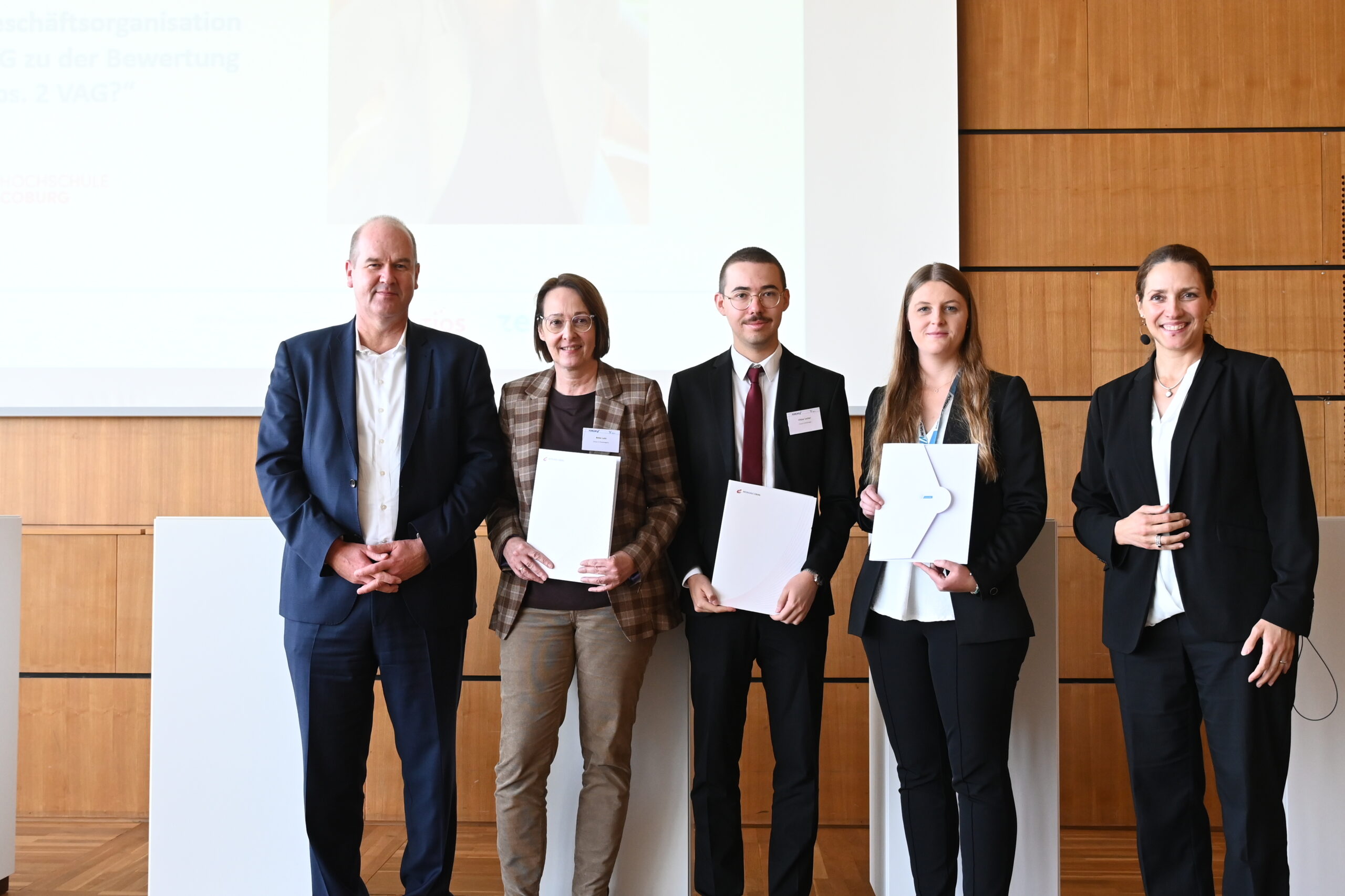 Eine Gruppe von fünf Personen steht in einem Raum der Hochschule Coburg. Drei halten Zertifikate oder Dokumente in den Händen, sind im Business-Outfit gekleidet und positionieren sich vor einer Holzkulisse und einem Bildschirm.