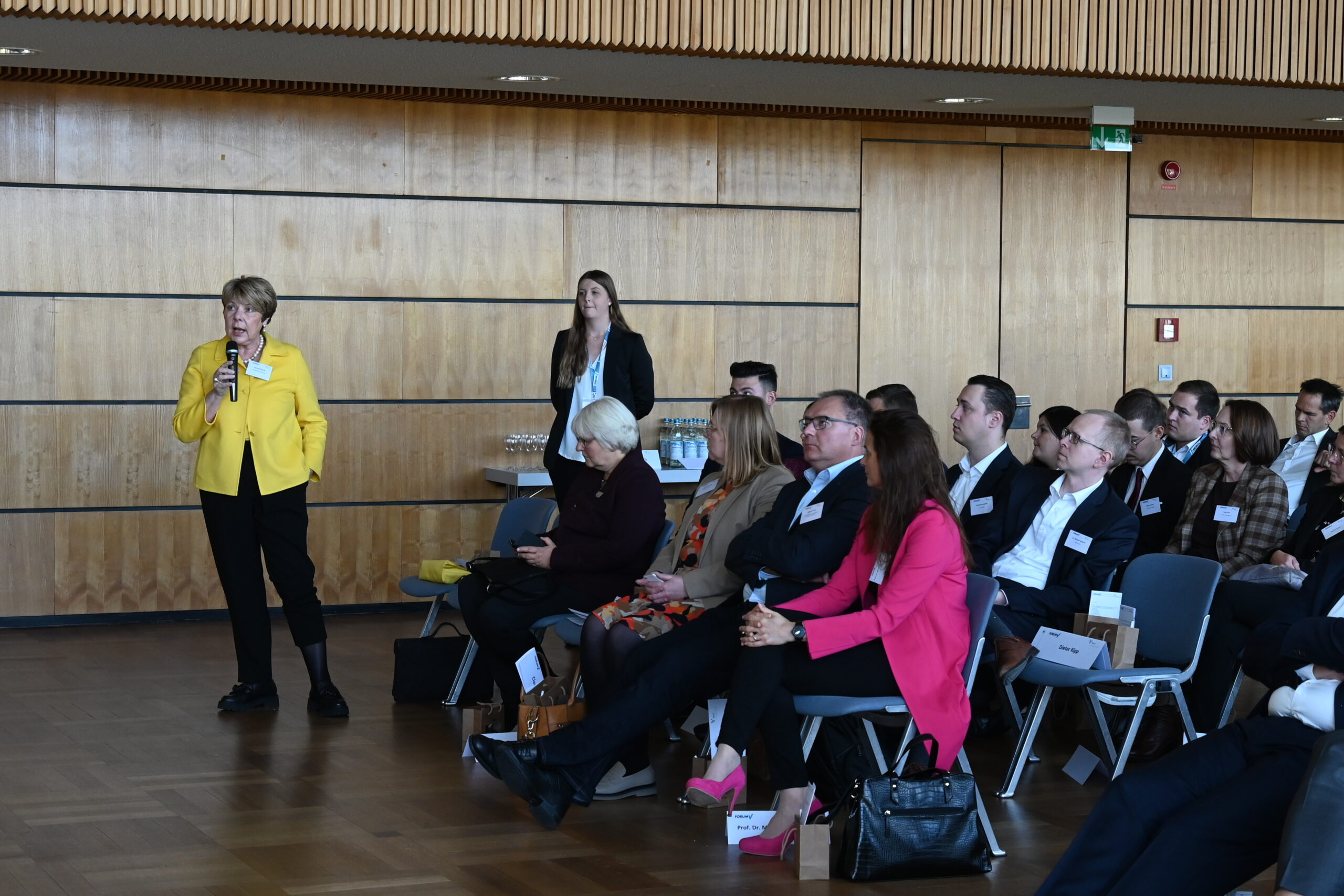 Eine Frau in einer gelben Jacke spricht in ein Mikrofon, während die Teilnehmer im Konferenzraum der Hochschule Coburg aufmerksam zuhören und einige sich Notizen machen. Hinter ihr bilden eine Holzwand und ein Tisch mit Gläsern die Kulisse für diese spannende Präsentation.