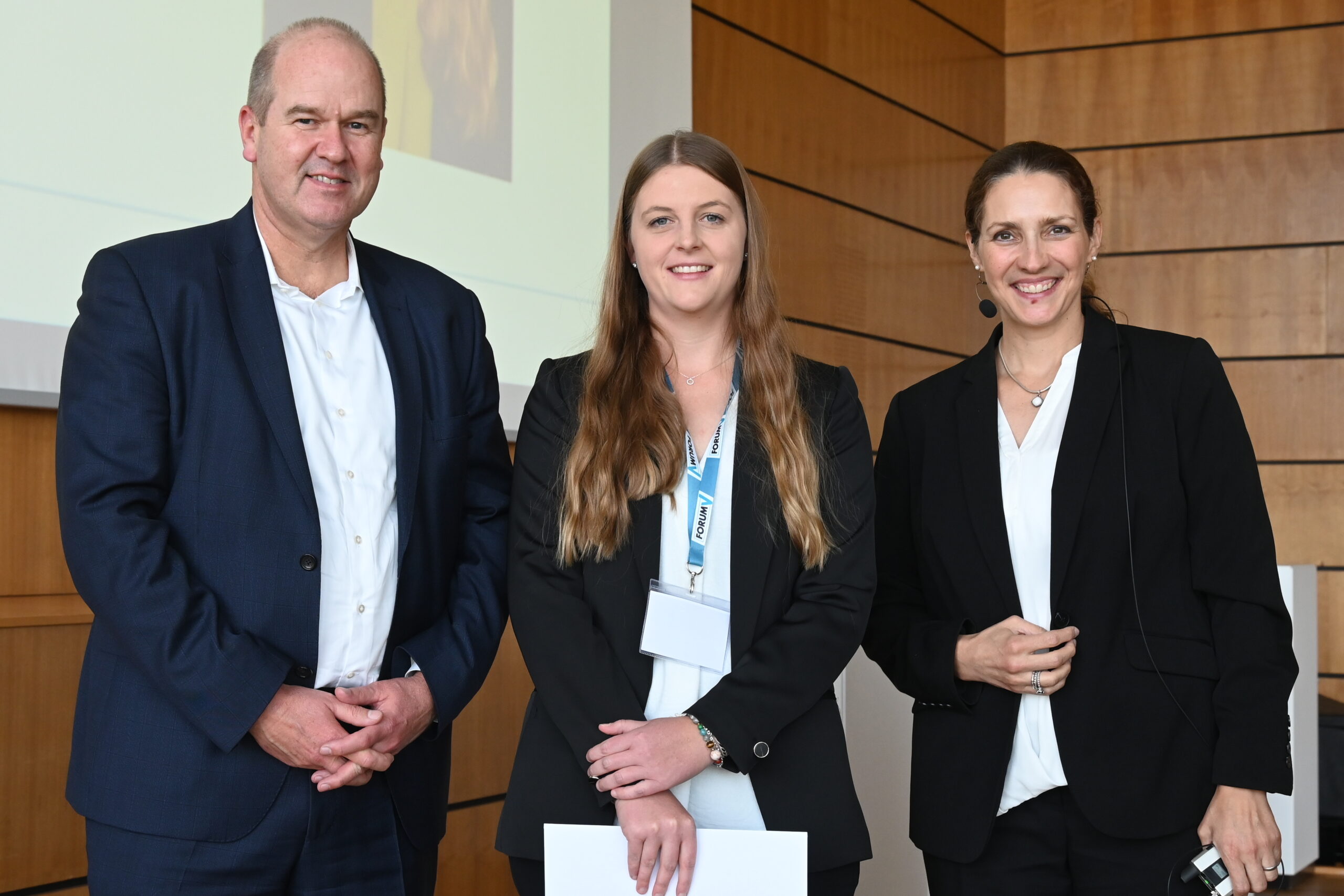 Drei Personen in Business-Kleidung stehen lächelnd in einem holzgetäfelten Raum zusammen, vermutlich bei einer Veranstaltung der Hochschule Coburg. Zwei Frauen und ein Mann halten Papiere in den Händen und tragen Namensschilder. Eine Frau hat ein Headset-Mikrofon, was darauf hindeutet, dass sie an einer professionellen Präsentation teilnehmen.