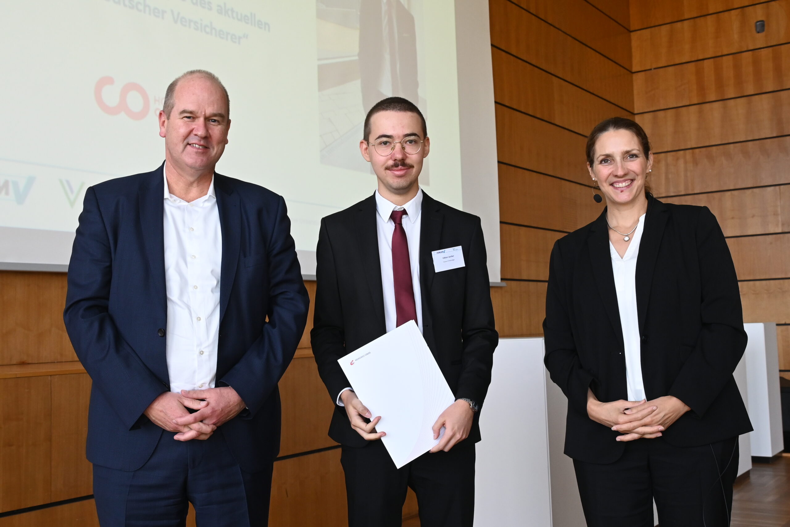 Drei Personen stehen drinnen und lächeln in die Kamera. Der Mann in der Mitte, der möglicherweise mit der Hochschule Coburg verbunden ist, hält einen weißen Ordner in der Hand. Sie sind in formelle Kleidung gekleidet, was auf eine berufliche Veranstaltung oder eine Preisverleihung hindeutet. Hinter ihnen ist eine Holzwand und ein Teilblick auf einen Bildschirm.