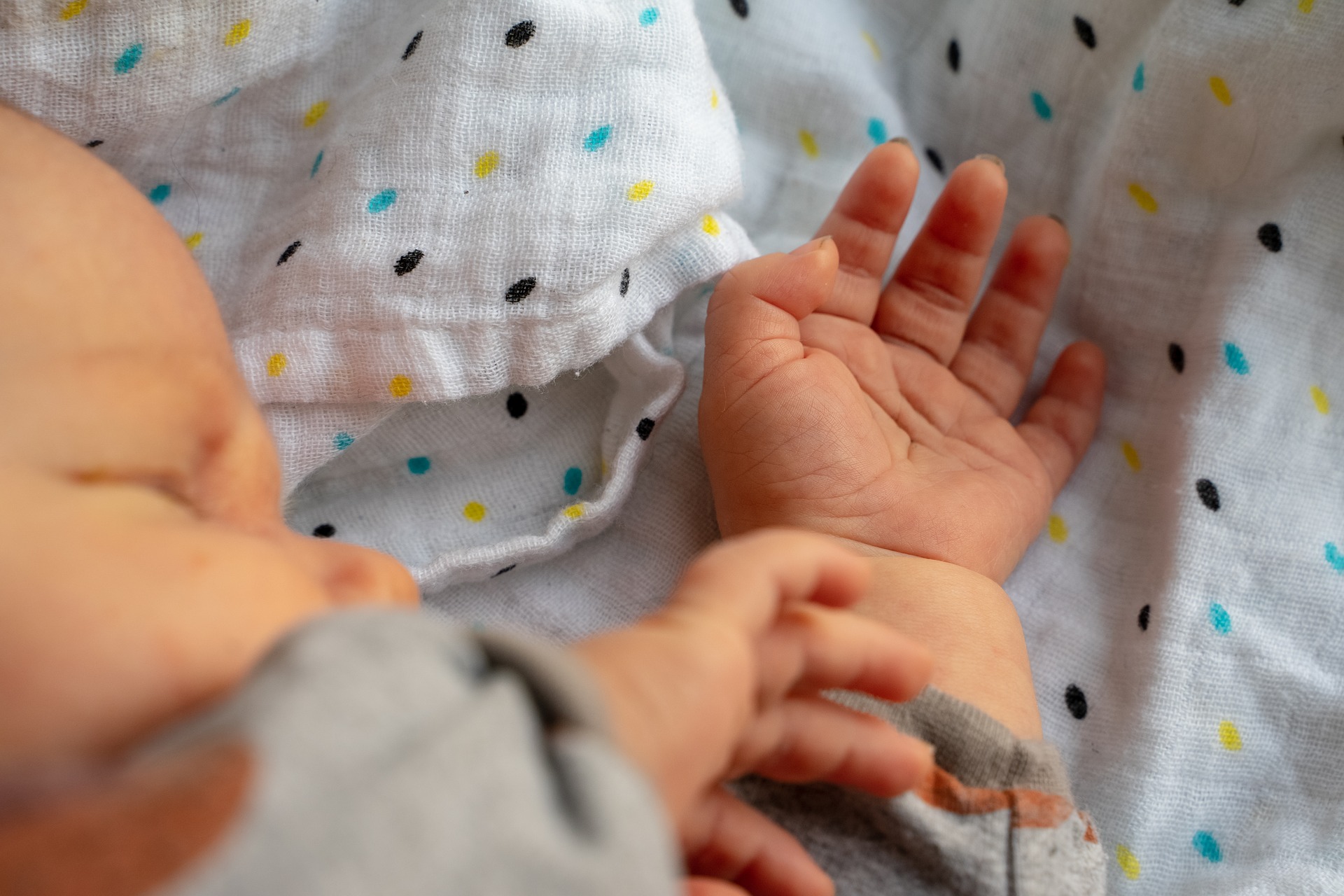 Ein Baby liegt auf einer weißen, gepunkteten Decke, eine Hand ist geöffnet, die andere leicht angehoben. Dies erinnert an eine heitere Szene aus einem Designkurs an der Hochschule Coburg. Das Gesicht des Babys kommt sanft hervor und wird von sanftem Licht angestrahlt.