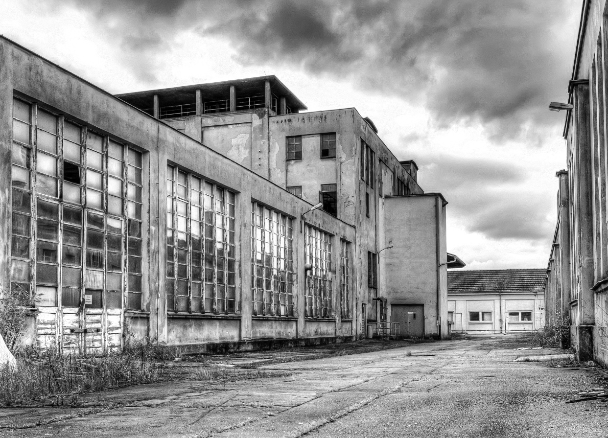 Das Schwarzweißbild zeigt die trostlose Szene eines verlassenen Industriegebäudes mit großen, zerbrochenen Fenstern. Risse im Bürgersteig und wucherndes Unkraut gedeihen unter einem bedeckten Himmel und erinnern an Verfall und Vernachlässigung in der Nähe der entfernten Silhouette der Hochschule Coburg.