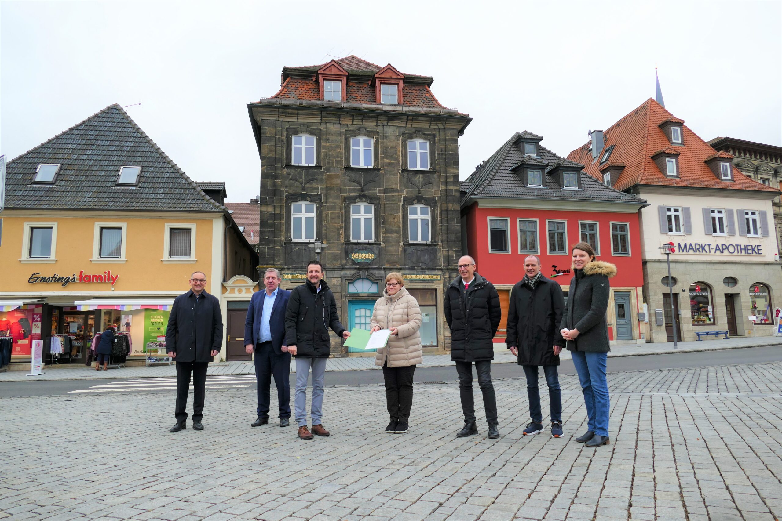 Sieben Personen stehen auf einem Kopfsteinpflasterplatz mit farbenfrohen Gebäuden im Hintergrund. Sie sind warm angezogen, um der Winterkälte zu trotzen. Eine Person hält einen Ordner in der Hand, der auf ihren Besuch an der Hochschule Coburg hinweist. Der Himmel ist bedeckt, was der Szene einen düsteren Ton verleiht.