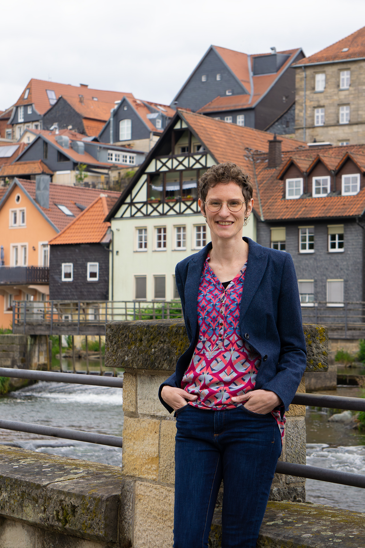 A person with short hair and glasses is smiling while standing on a bridge near Hochschule Coburg. They are wearing a blue blazer and a colorful patterned blouse. The background features a river and several colorful, half-timbered houses with orange roofs.