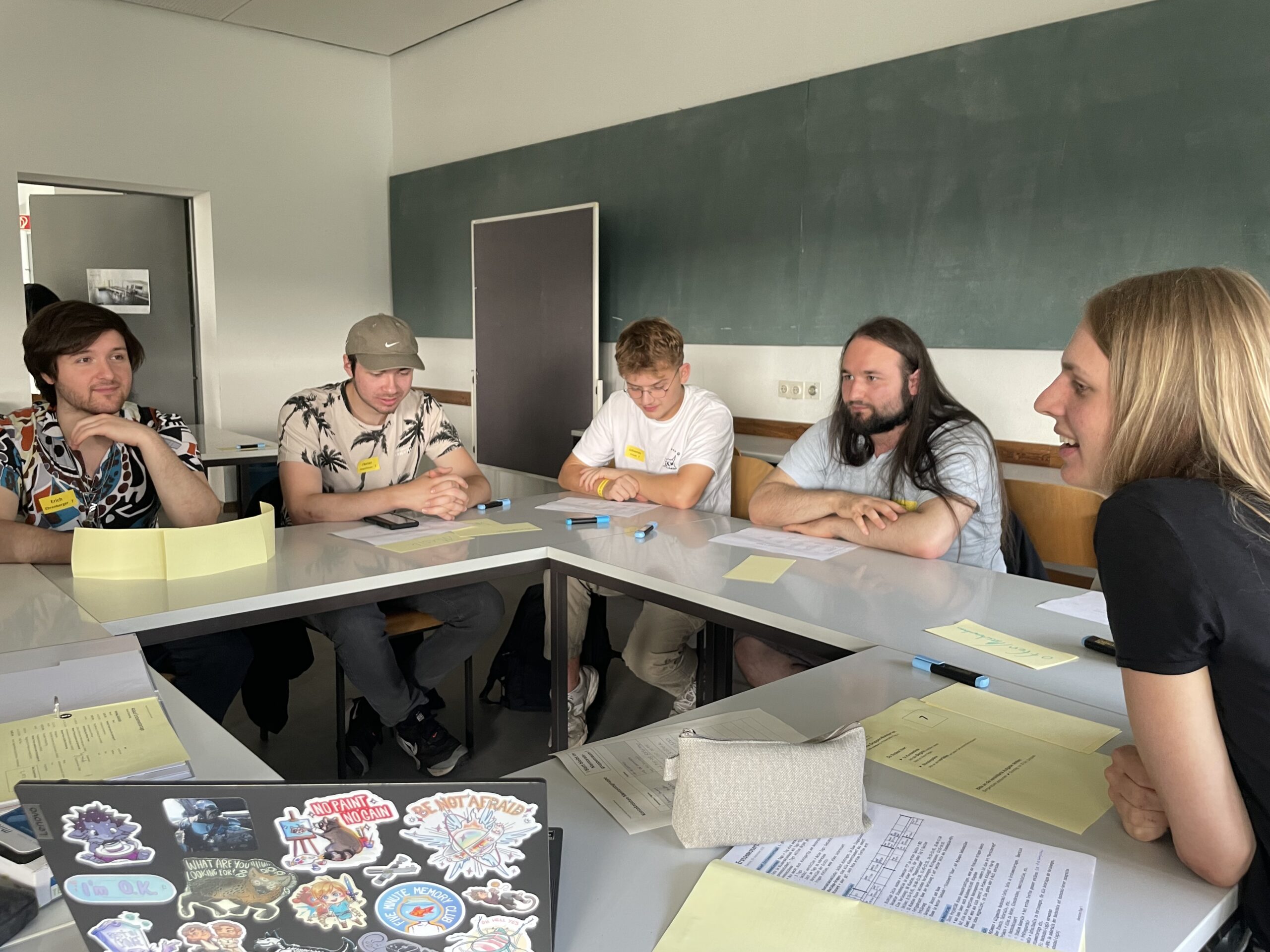 Fünf Personen sitzen an einem Tisch in der Hochschule Coburg und diskutieren vertieft. Papiere, Stifte und ein mit bunten Aufklebern verzierter Laptop liegen verstreut auf dem Tisch. Eine große Tafel und eine Tür bilden den Hintergrund für diesen lebhaften akademischen Austausch.