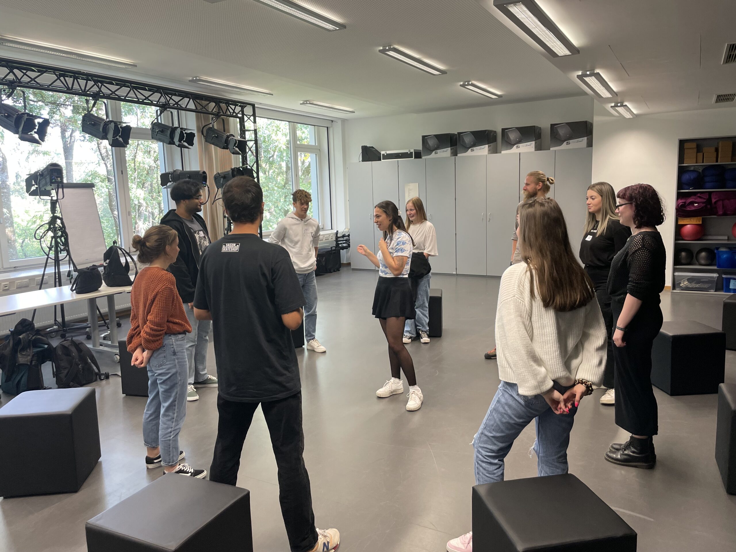 Eine Gruppe von Menschen steht in einem Kreis unter Studiobeleuchtung an der Hochschule Coburg. Der Raum ist in eine lebhafte Diskussion oder Aktivität vertieft und verfügt über große Fenster, ein Whiteboard und verschiedene Geräte, die ordentlich auf Regalen angeordnet sind.