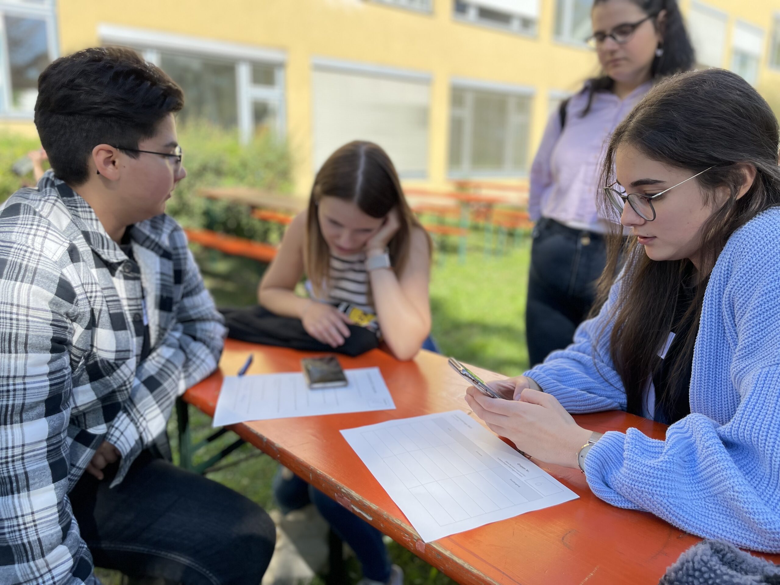 Vier Personen sitzen um einen orangefarbenen Picknicktisch im Freien an der Hochschule Coburg. Zwei Frauen konzentrieren sich auf ihre Telefone, eine blickt über ihre Unterlagen und ein Mann hört aufmerksam zu. Sie scheinen gemeinsam an einem Projekt oder einer Lernsitzung zu arbeiten.