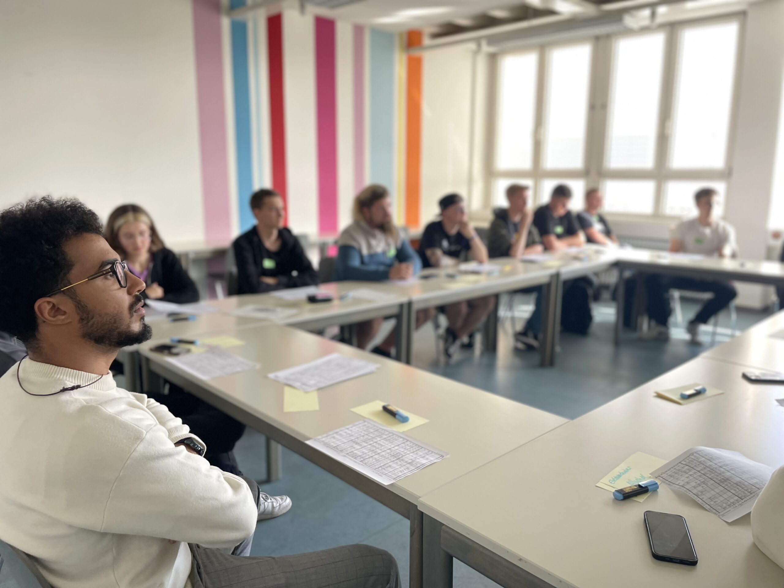 In einem Hörsaal der Hochschule Coburg mit bunt gestreiften Wänden sitzt eine Gruppe um Tische herum und hört einem Vortragenden aufmerksam zu. Papier und Stifte liegen verstreut herum, während das Sonnenlicht durch das große Fenster fällt und eine Person auf der linken Seite in den Mittelpunkt rückt.