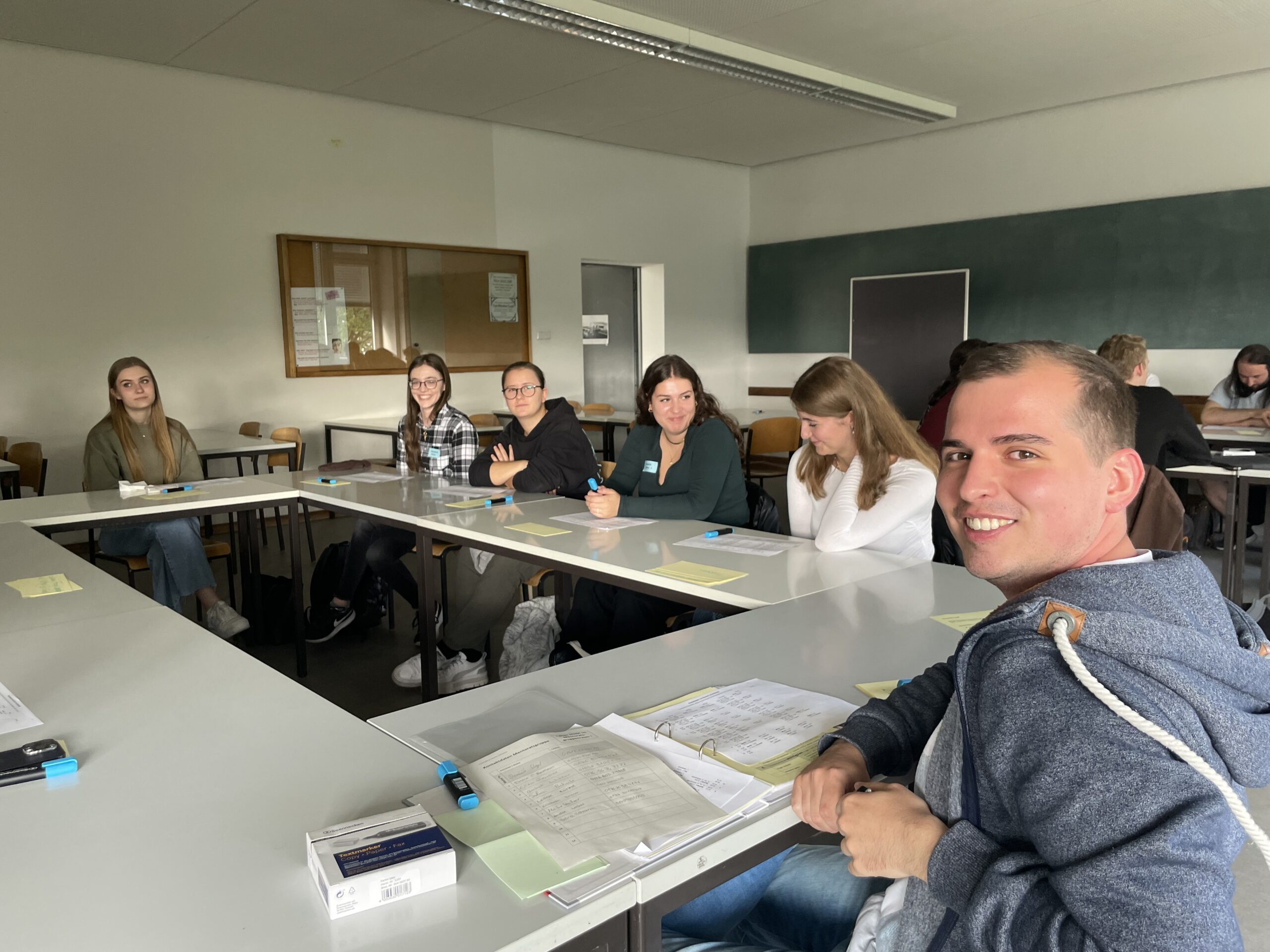 Eine Gruppe von Menschen sitzt in einem Hörsaal der Hochschule Coburg an U-förmig angeordneten Tischen. Sie wirken engagiert und aufmerksam, vor ihnen liegen Notizbücher und Stifte. Der Raum verfügt über eine Tafel und ein schwarzes Brett, und durch das Fenster fällt natürliches Licht in den Raum.