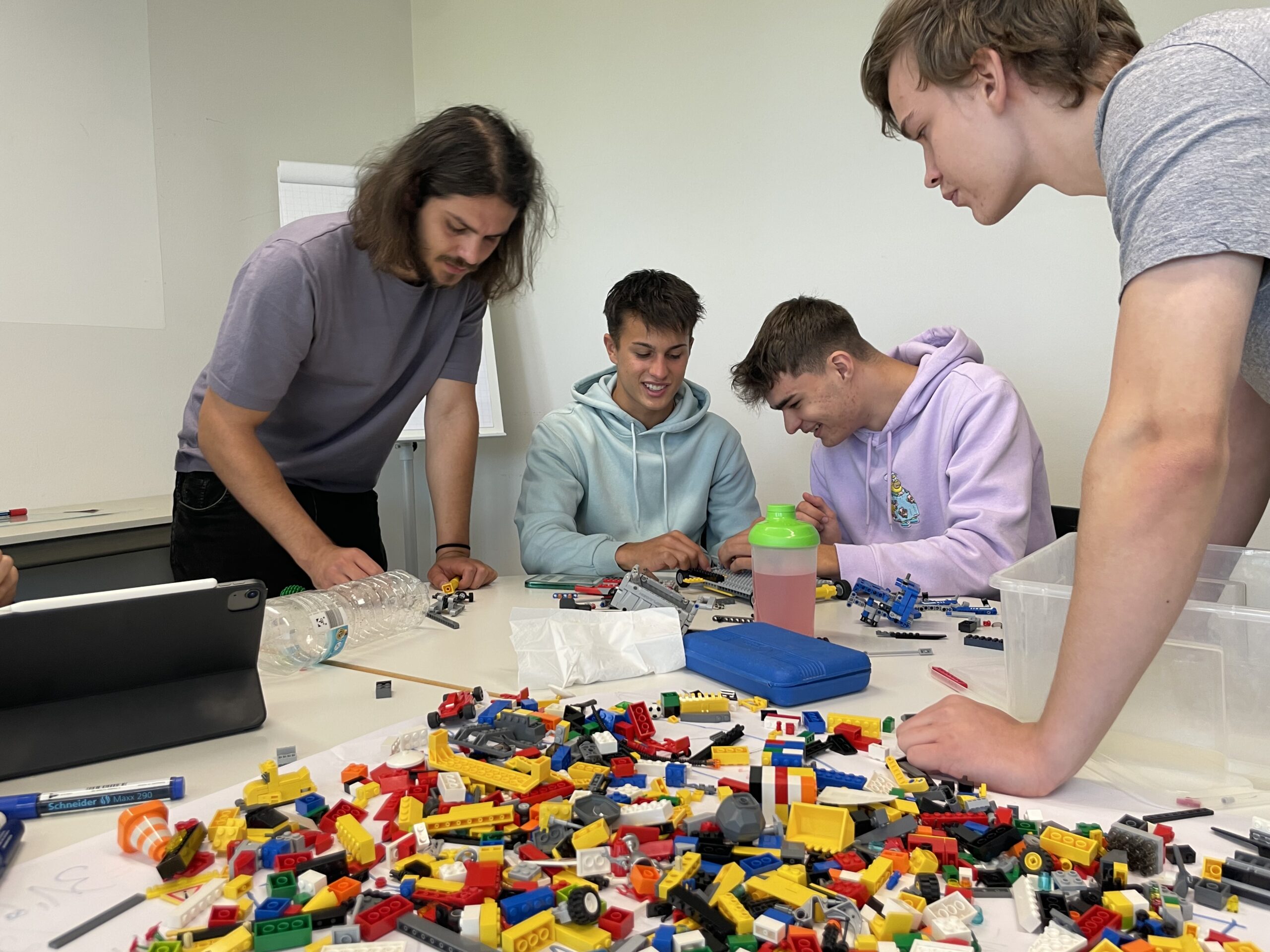 Vier Personen sitzen an einem mit bunten LEGO-Steinen bedeckten Tisch in der Hochschule Coburg. Sie scheinen einer gemeinsamen Tätigkeit nachzugehen und sich darauf zu konzentrieren, gemeinsam etwas zu bauen. Im Raum herrscht eine lockere und kreative Atmosphäre.