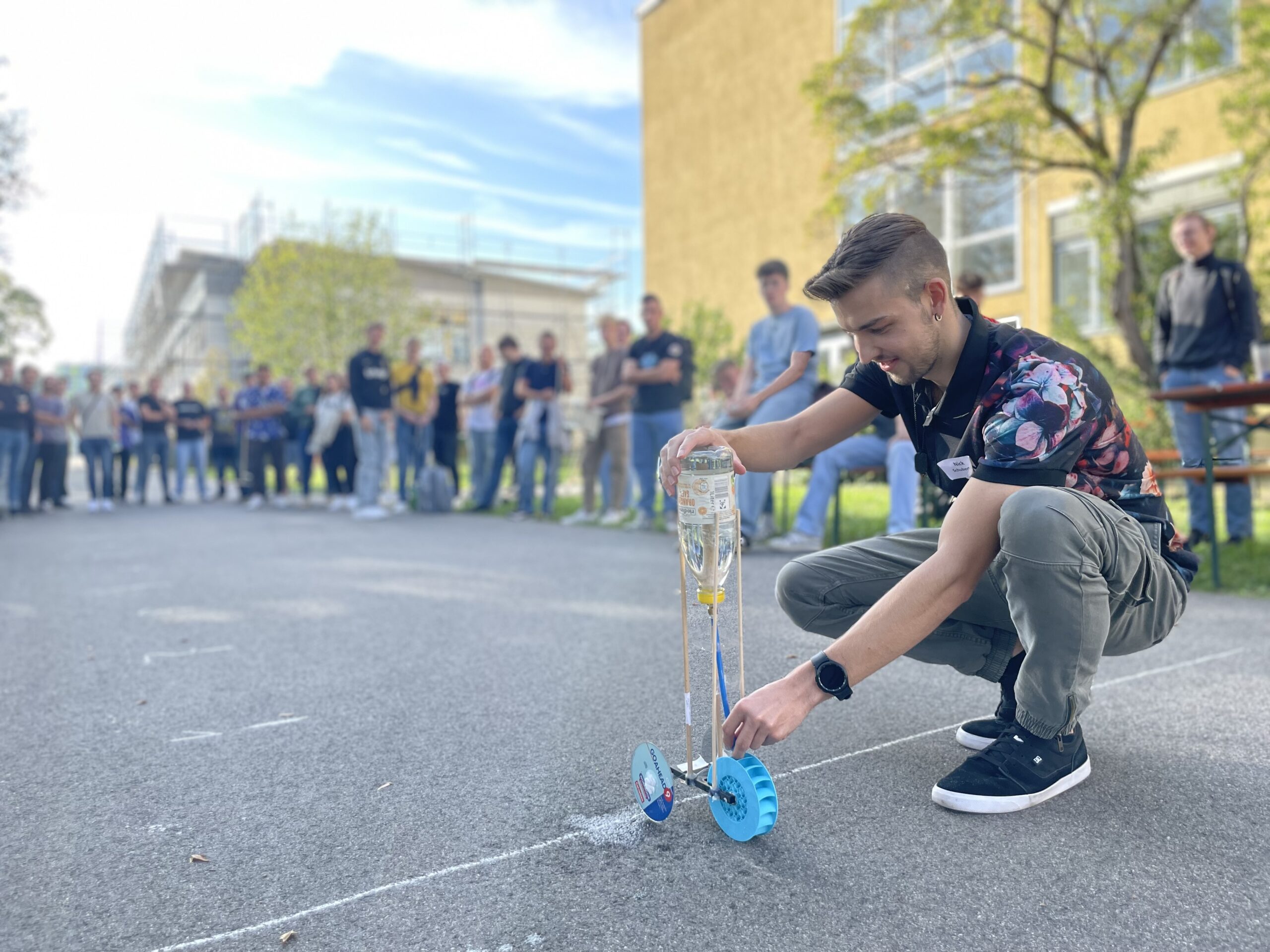 Ein junger Mann hockt auf einer gepflasterten Fläche und hält ein kleines Fahrzeug mit Rädern aus einer Plastikflasche auf dem Campus der Hochschule Coburg. Er ist von Zuschauern umgeben, in einer entspannten Außenumgebung mit Bäumen und Gebäuden im Hintergrund.