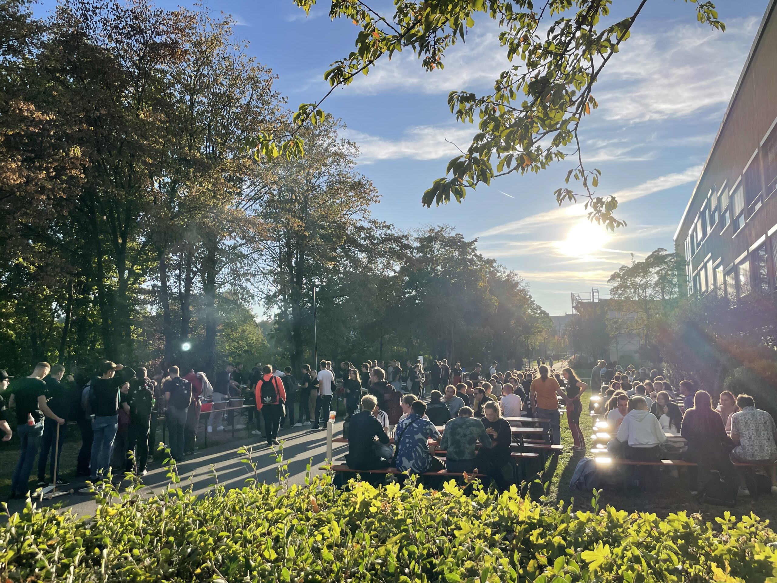 Eine große Gruppe von Menschen, darunter auch Studierende der Hochschule Coburg, versammelt sich draußen in einer parkähnlichen Umgebung mit Tischen unter dem sonnigen Himmel. Üppige Bäume und Büsche umgeben den Bereich, und alle genießen das angenehme Wetter.