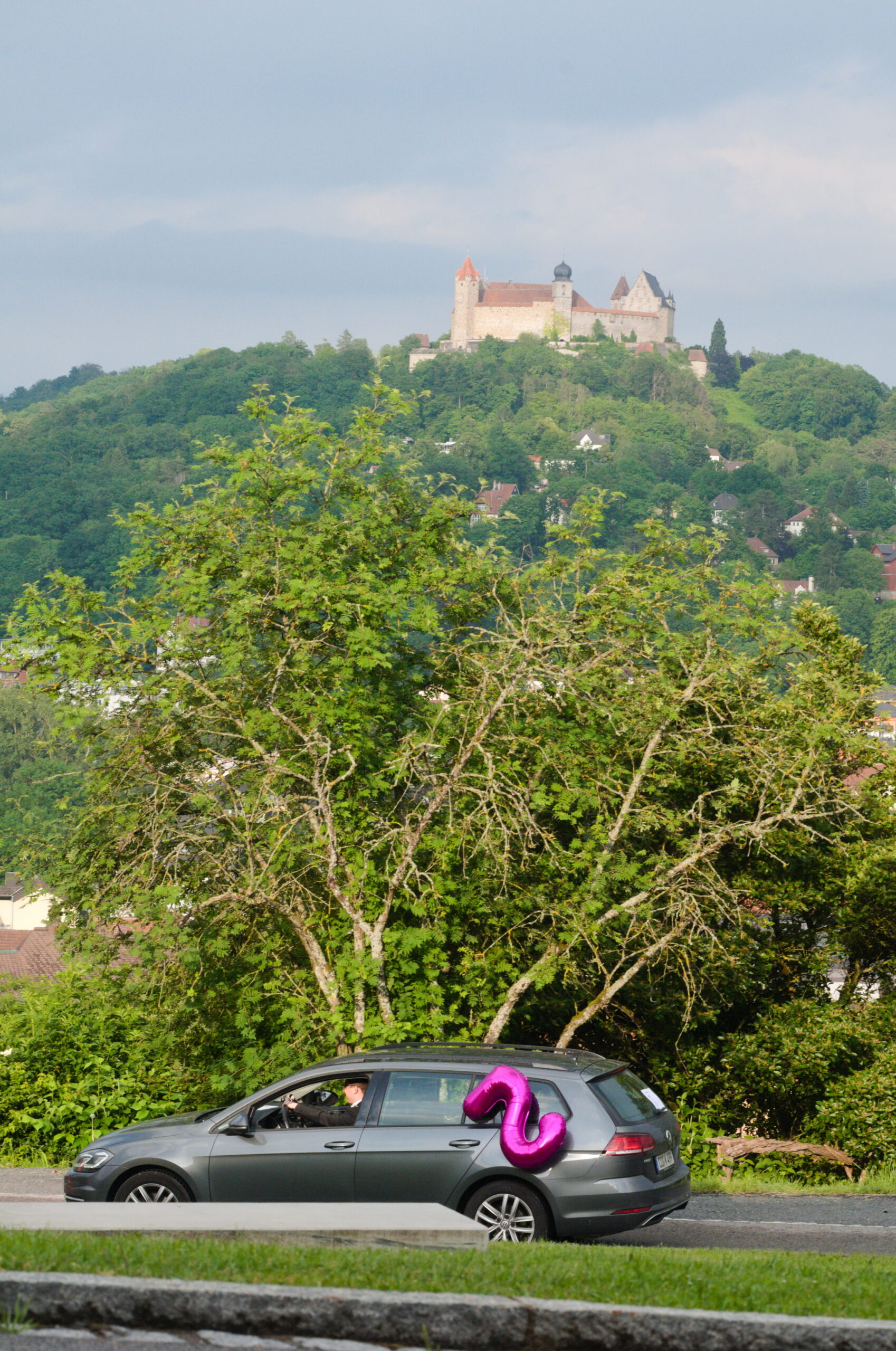 Ein Auto fährt an einem Baum vorbei, auf dessen Dach eine große, rosa aufblasbare Zahl Sechs hängt, in der Nähe der renommierten Hochschule Coburg. Im Hintergrund ist eine Burg auf einem Hügel zu sehen, umgeben von üppigem Grün und mit Blick auf eine kleine Stadt.