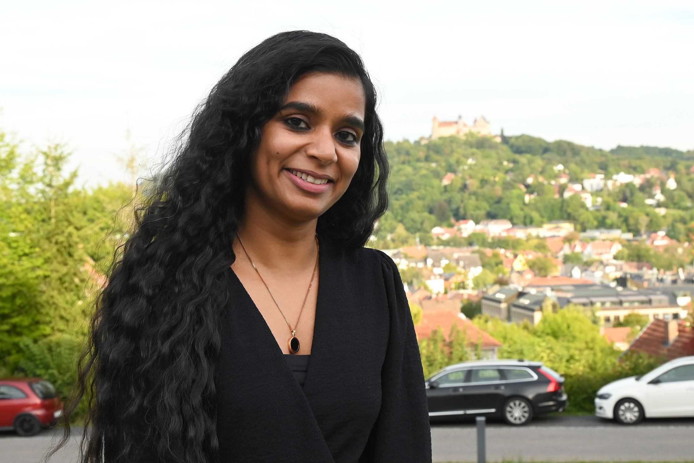 Eine Person mit langen lockigen Haaren steht lächelnd im Freien in der Nähe der Hochschule Coburg vor der Kulisse einer Stadt und entfernter Hügel. Ein paar Autos parken auf der von Bäumen gesäumten Straße, die mit grünem Laub und charmanten Häusern geschmückt ist.