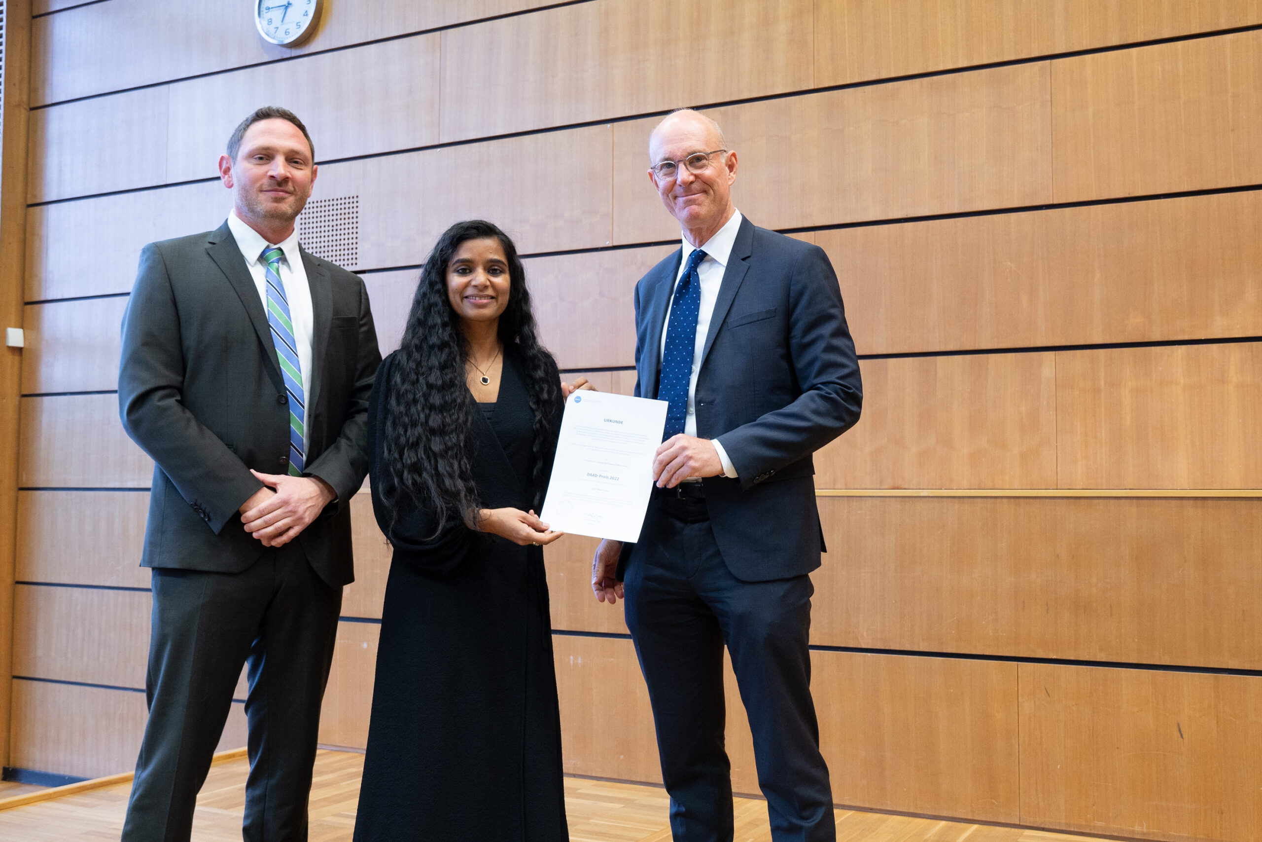 Drei Personen in formeller Kleidung stehen drinnen vor einer Holzwand in der Hochschule Coburg. Eine Frau in der Mitte hält ein Zertifikat, während zu beiden Seiten zwei Männer lächeln. Über ihnen ist eine Uhr zu sehen.