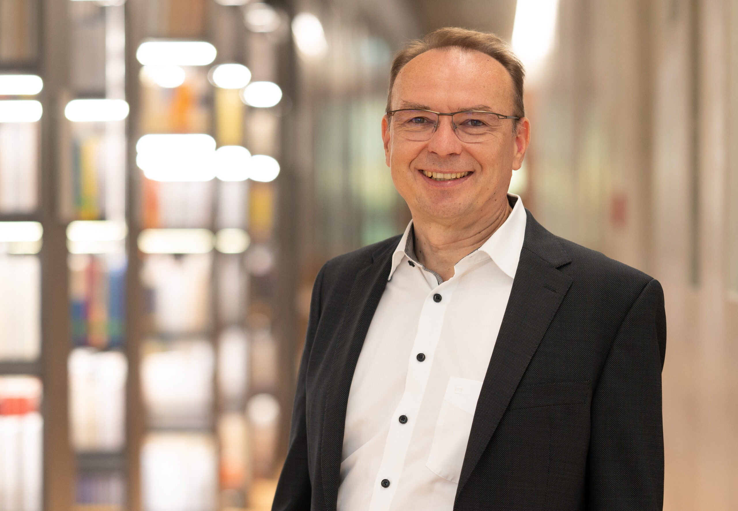 Ein Mann mit Brille, weißem Hemd und dunklem Blazer lächelt in die Kamera. Er steht in einem Innenbereich der Hochschule Coburg und zeigt vor einem unscharfen Hintergrund Bücherregale und moderne Beleuchtung.