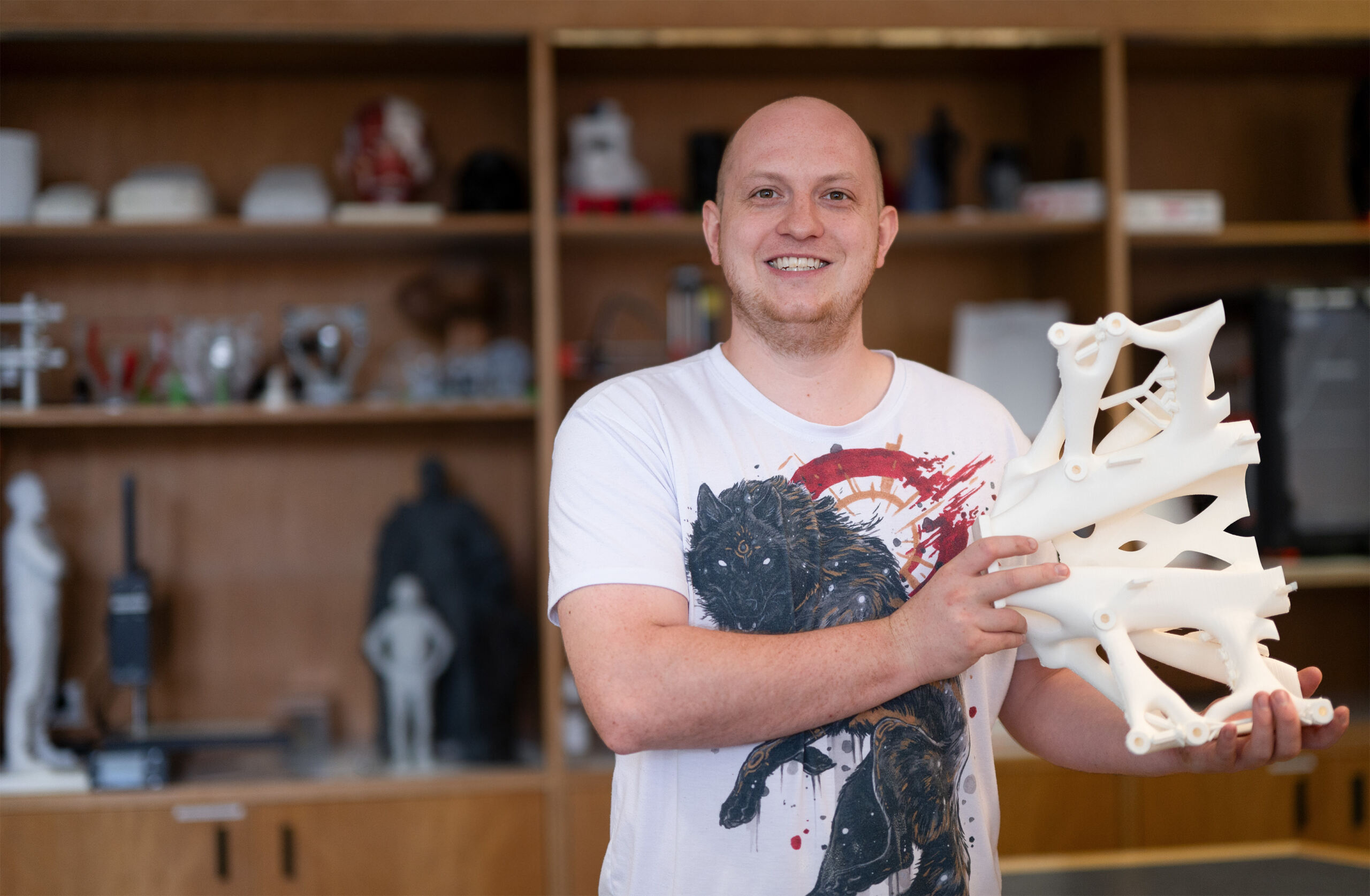 A person smiles while holding a complex 3D-printed structure, proudly standing in a room at Hochschule Coburg. Wooden shelves display various innovative 3D models and objects. The person wears a white t-shirt adorned with a vibrant graphic design, reflecting their creative spirit.