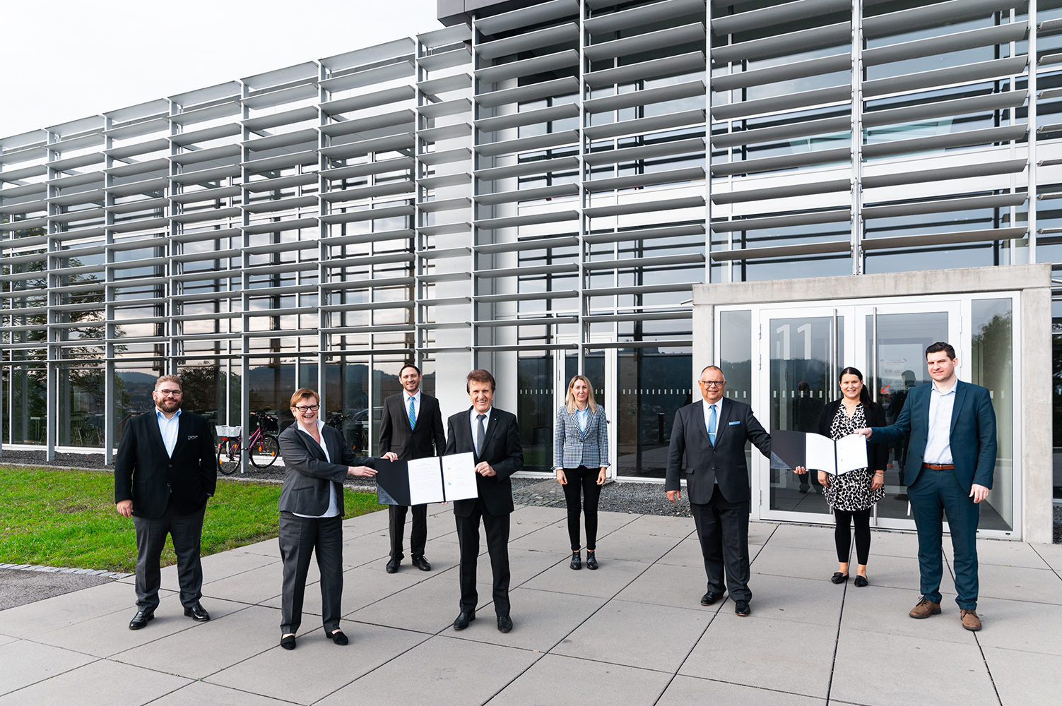 Sieben Personen in Business-Kleidung stehen vor einem modernen Gebäude der Hochschule Coburg mit Metallverkleidung. Drei Personen halten Zertifikate in der Hand, stehen etwas abseits, während andere lächeln. Die Szene lässt auf eine berufliche oder feierliche Veranstaltung schließen.