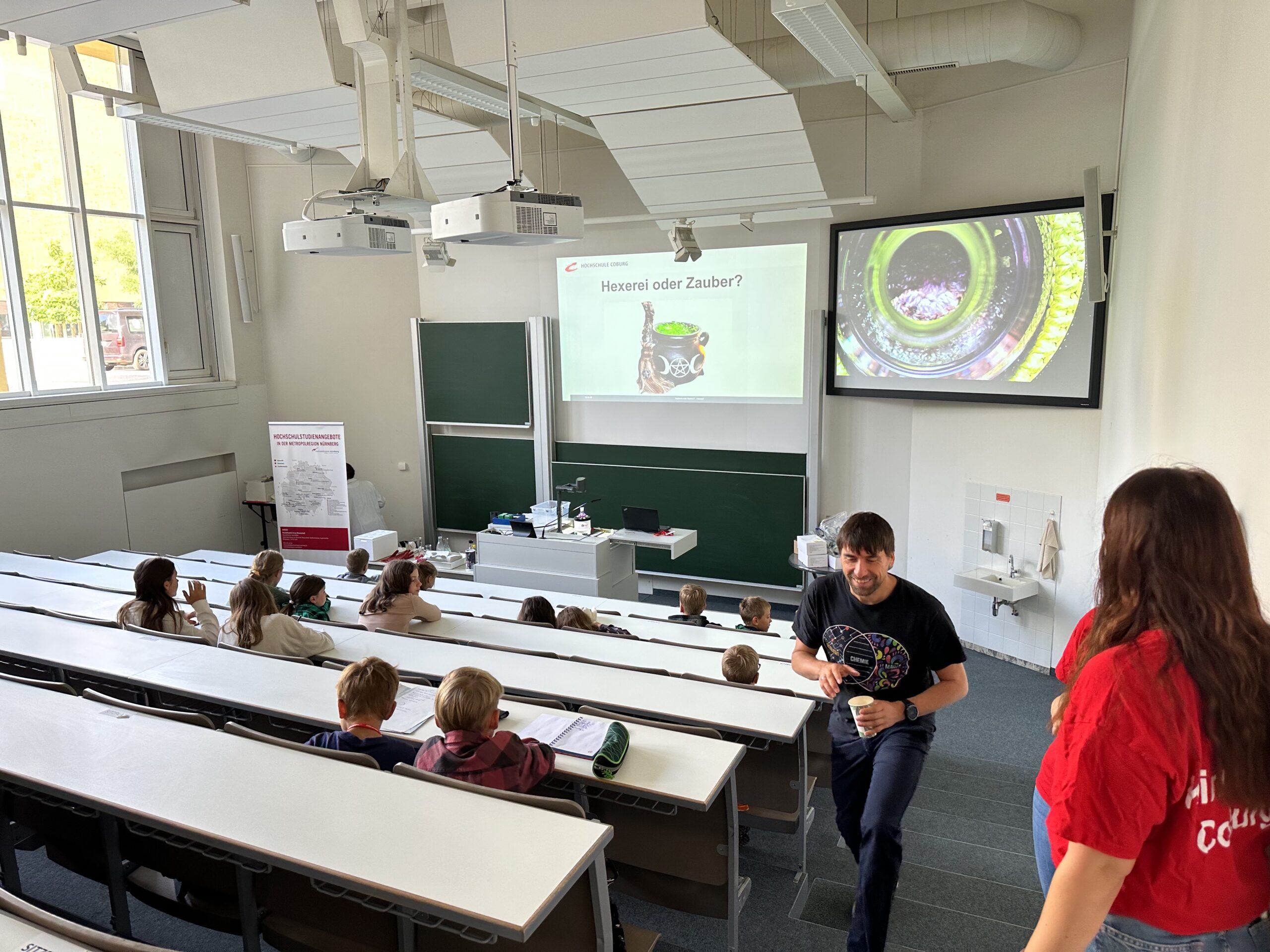 In einem Hörsaal der Hochschule Coburg sitzen die Studenten in abgestuften Reihen, ihre Augen sind auf den Dozenten und die Präsentation gerichtet. Große Fenster erhellen die weißen Wände, während auf einer Projektionsfläche ein Kameraobjektivbild mit deutschem Text angezeigt wird. Zwei Personen stehen in der ersten Reihe und diskutieren.