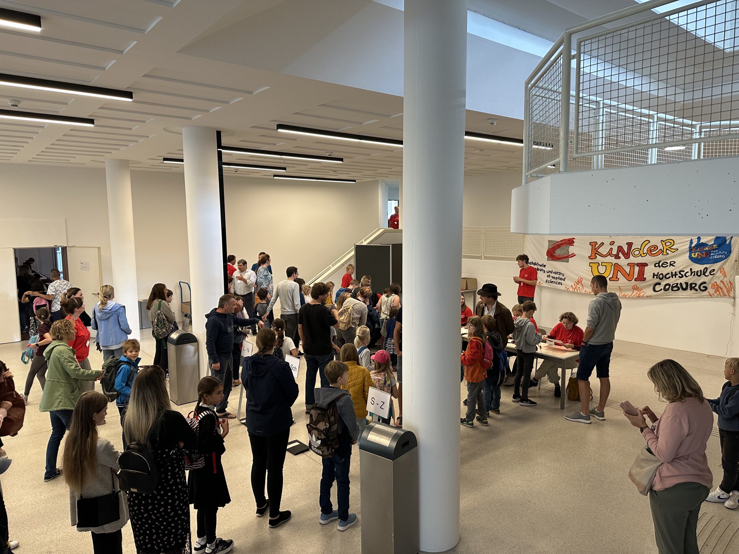 Eine große Gruppe versammelt sich in einem Gebäude; Kinder mit Rucksäcken und Erwachsene mischen sich untereinander. Auf einem Banner prangt stolz „Kinder UNI“ neben „Hochschule Coburg“. Einige Leute sitzen an Tischen und genießen die lebhafte Atmosphäre, andere stehen und unterhalten sich.