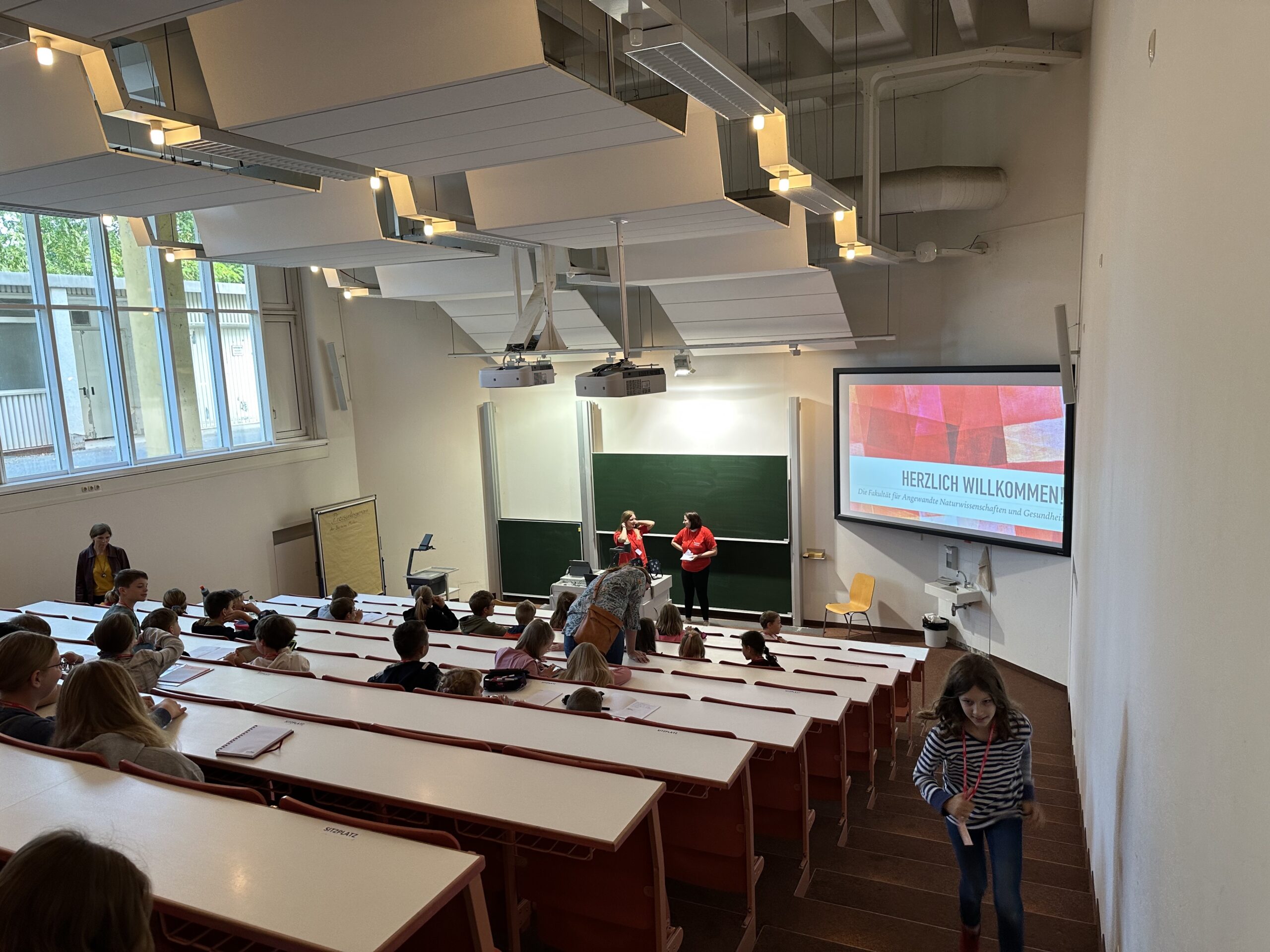 In einem Hörsaal der Hochschule Coburg sitzen Studierende vor einer großen Leinwand mit der Aufschrift „Herzlich Willkommen“. Vorne stehen zwei Vortragende neben einer Tafel. Der Raum ist mit Sitzreihen ausgestattet und hat auf einer Seite große Fenster.