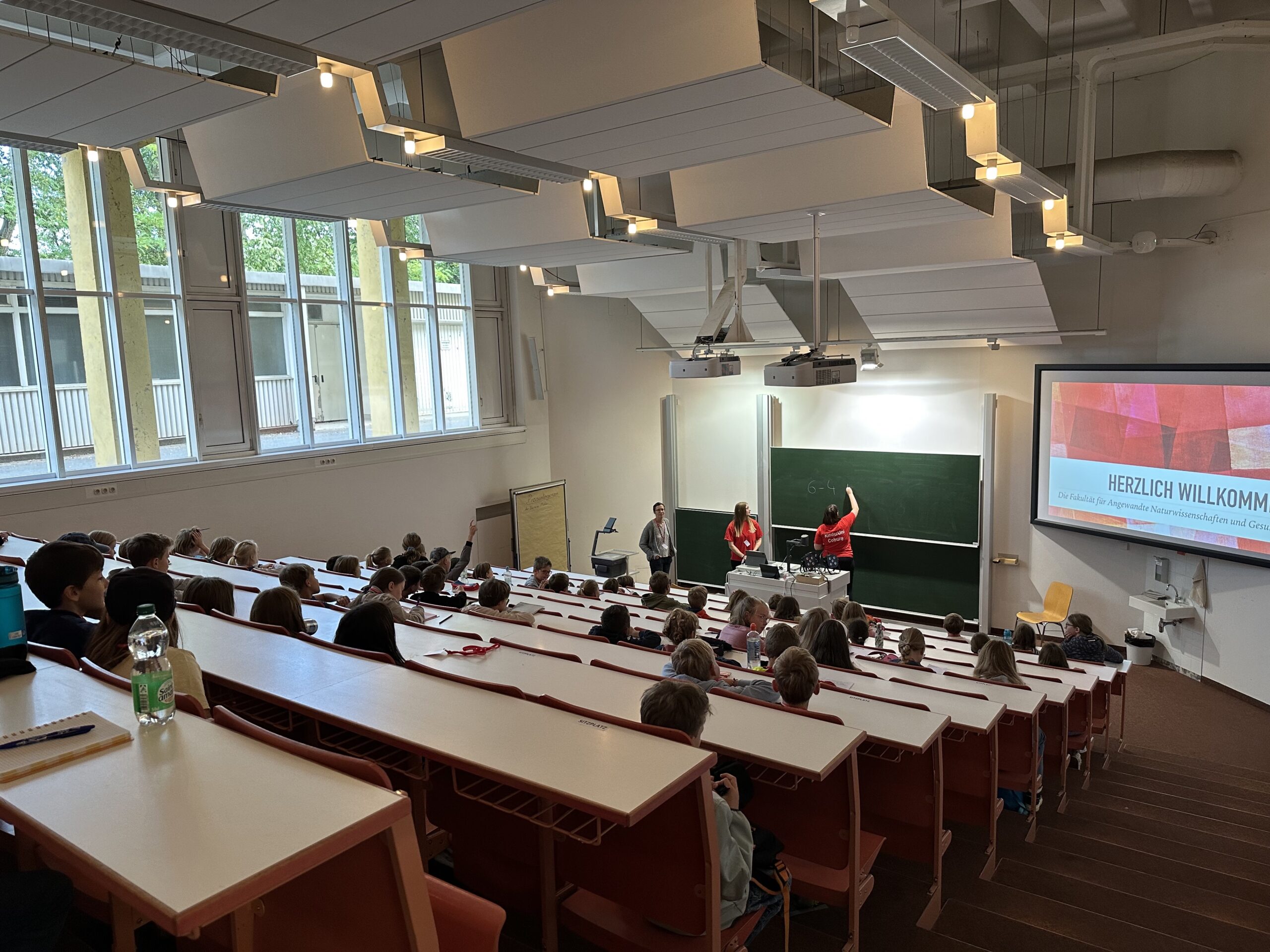 Ein Hörsaal der Hochschule Coburg ist voller Studenten, die an ihren Schreibtischen sitzen und neben einer Tafel und einer Projektionsleinwand einer Podiumsdiskussion gegenübersitzen. Auf der Leinwand steht "Herzlich Willkommen!", und große Fenster lassen Tageslicht in den Raum.