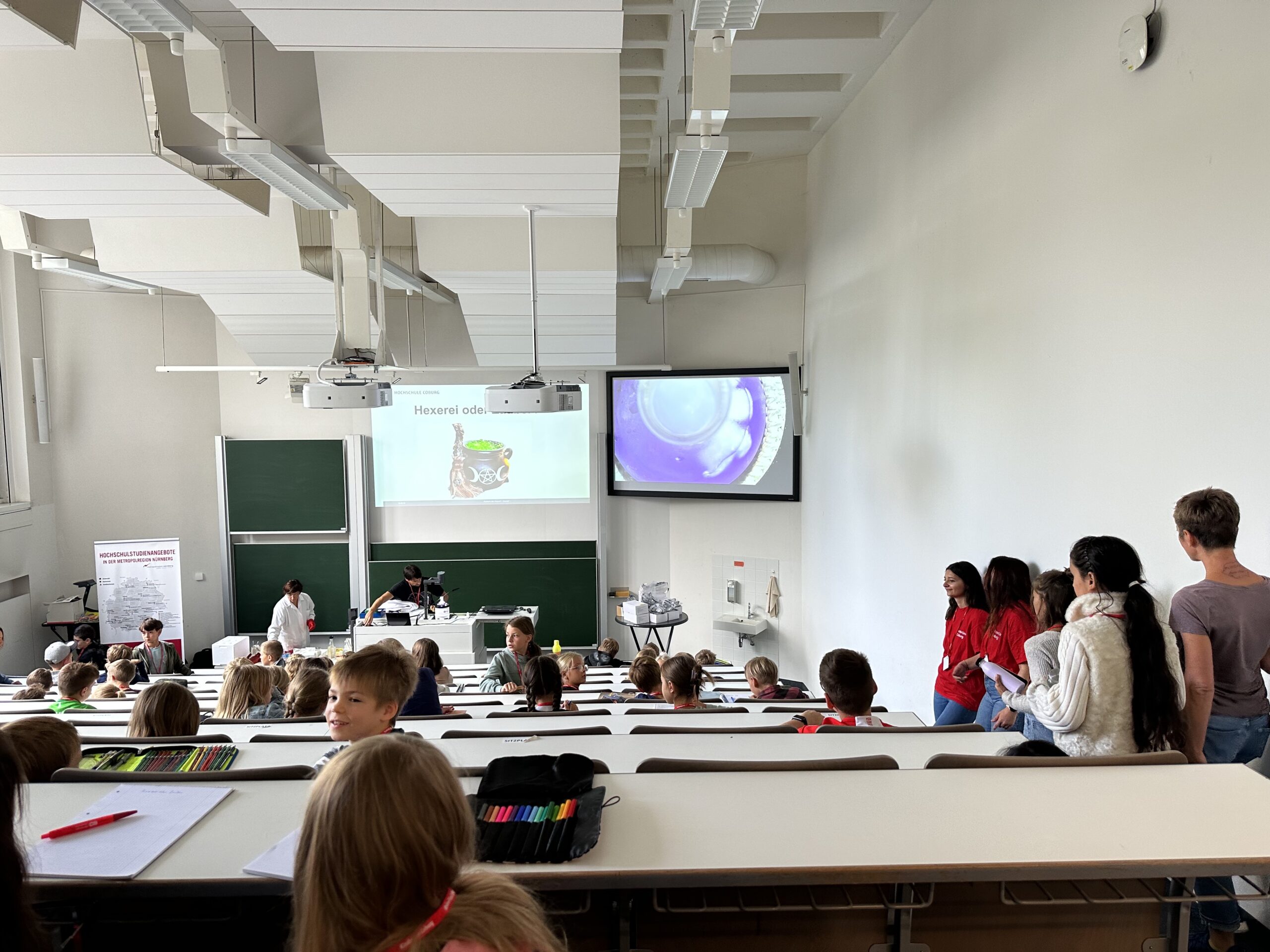 Studierende sitzen in einem Hörsaal der Hochschule Coburg, auf zwei Bildschirmen werden wissenschaftliche Bilder gezeigt. Vorne steht ein Dozent, an der Seite versammelt sich eine Gruppe in roten Hemden, die für lebendige akademische Atmosphäre sorgt.