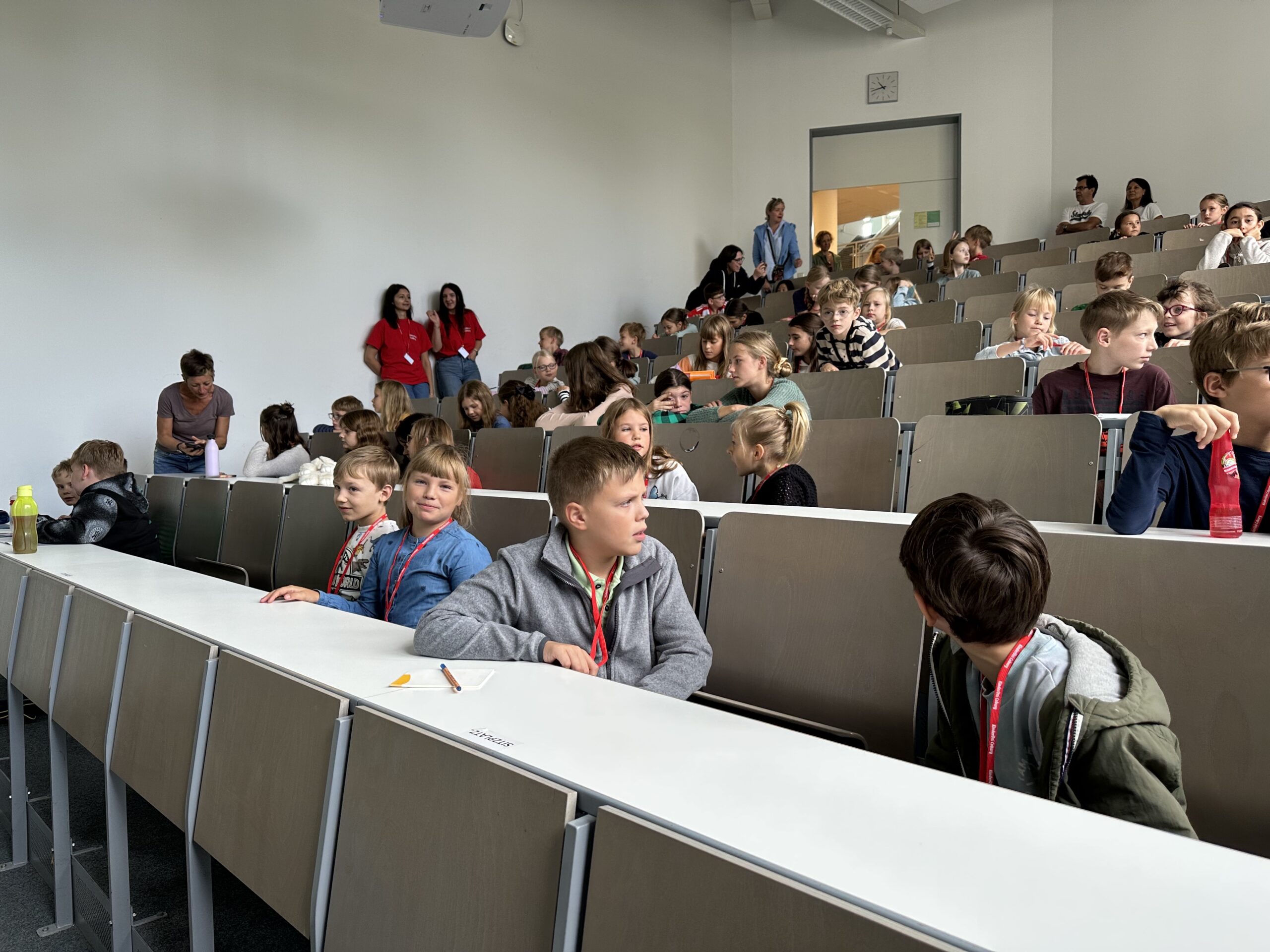 In einem gut beleuchteten Unterrichtsraum der Hochschule Coburg sitzen die Kinder in einer Sitzordnung wie im Hörsaal. Sie unterhalten sich lebhaft und blicken durch den Raum, in dem auch einige Erwachsene anwesend sind. Im Hintergrund ist eine weiße Tafel angebracht, die eine Atmosphäre des Lernens und Entdeckens fördert.