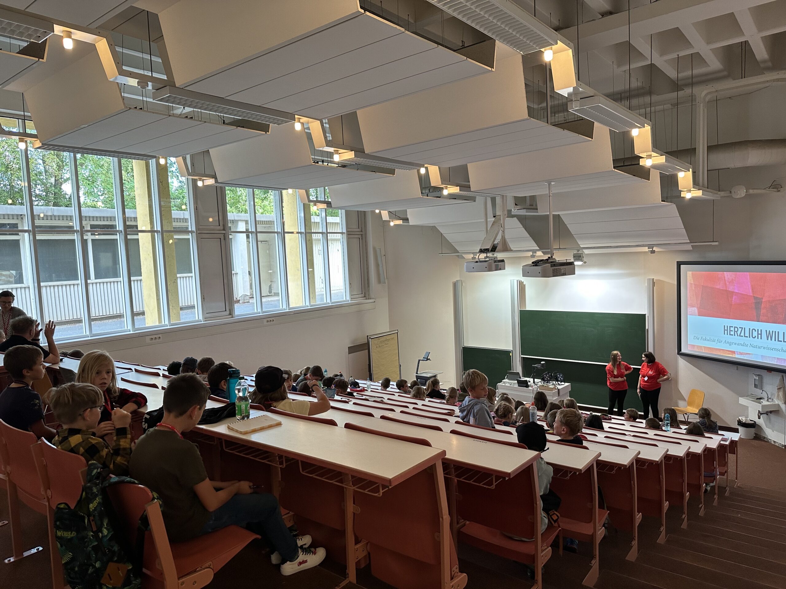 Ein Hörsaal der Hochschule Coburg ist voller Studenten, die in Reihen an Tischen sitzen und nach vorne blicken, wo zwei Vortragende neben einem großen Bildschirm stehen. Die hohen Decken und großen Fenster durchfluten den Raum mit natürlichem Licht.